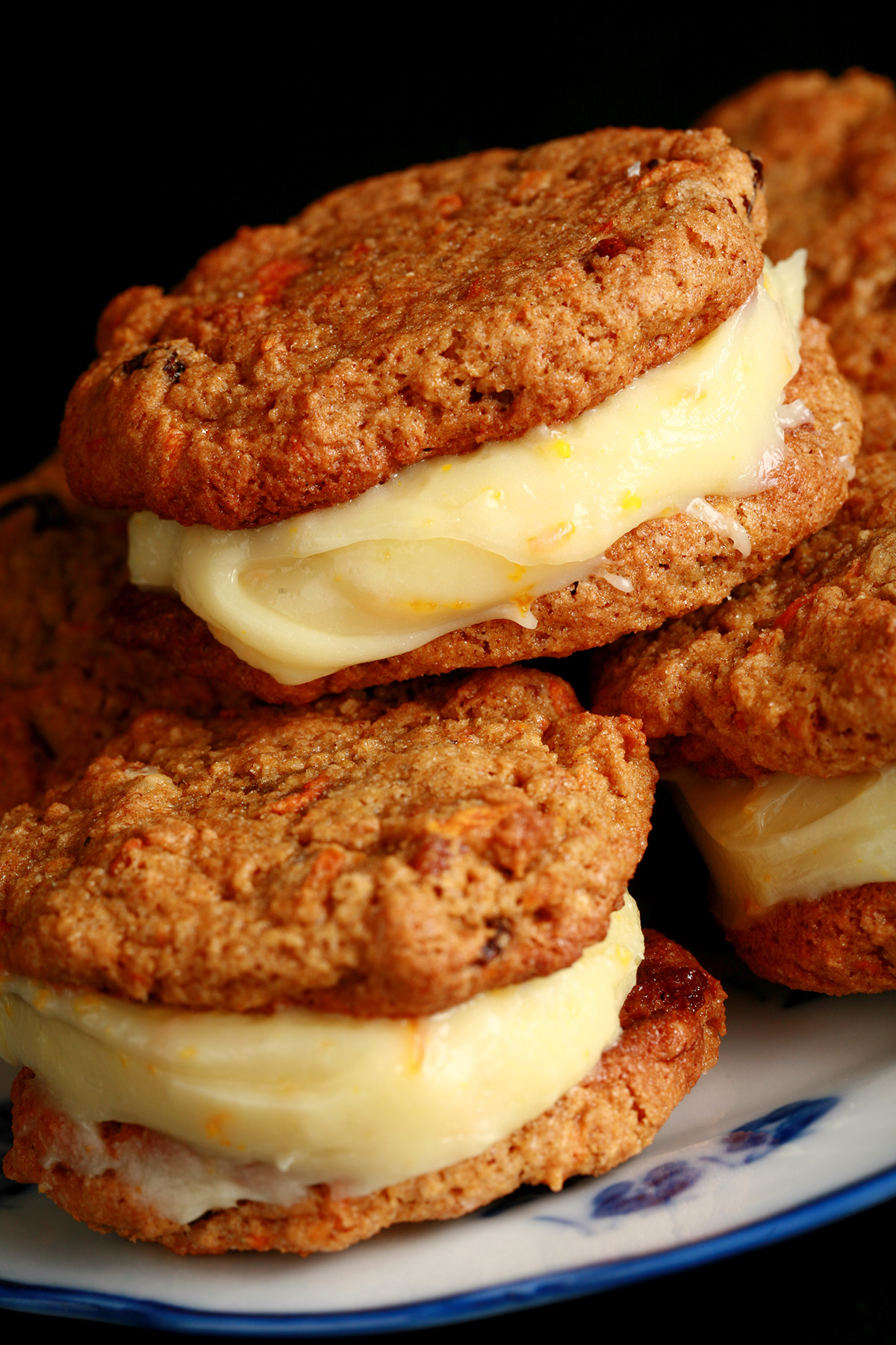 A small blue and white plate with a stack of carrot cake sandwich cookies on it. The filling is ivory coloured and has flecks of orange zest throughout.