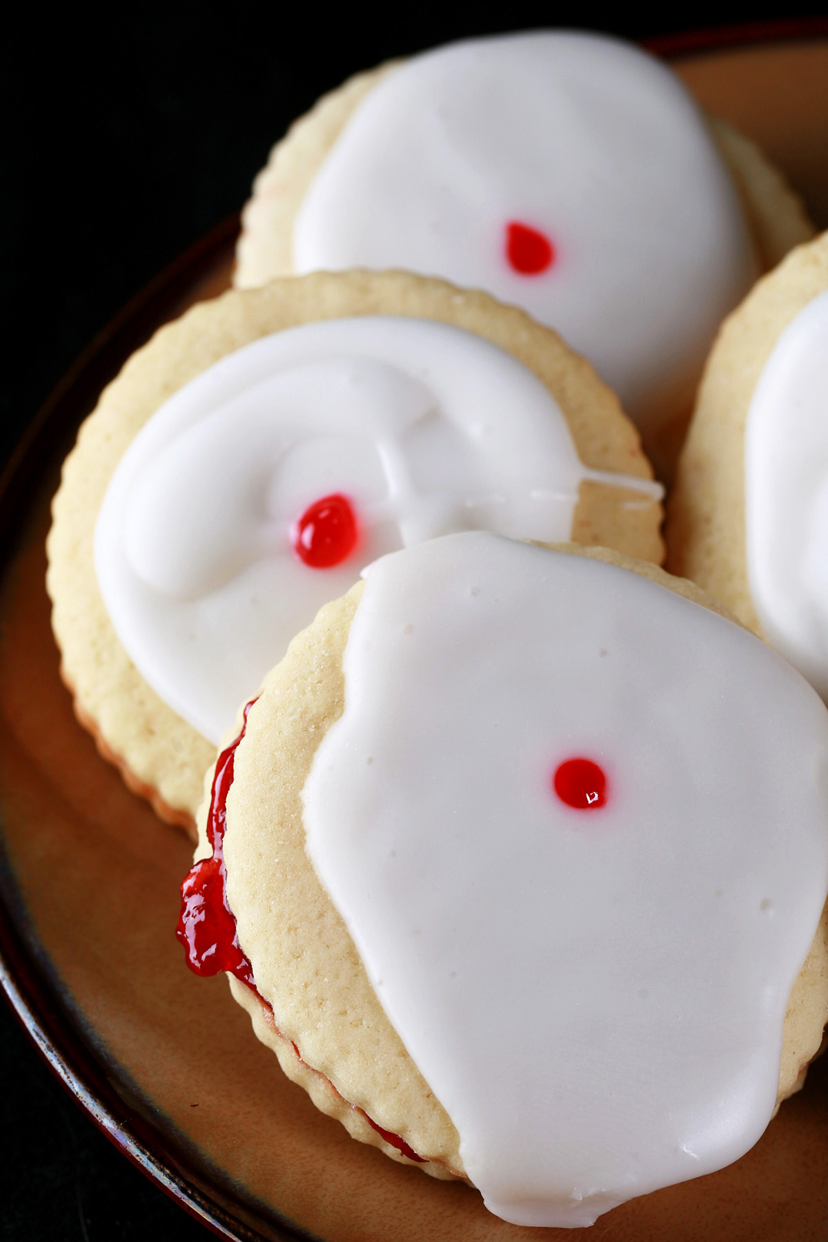 Close up view of a plate of gluten-free imperial cookies: Sandwich cookies filled with raspberry jam, frosted with a white glaze, and finished off with a dot of red gel in the center.
