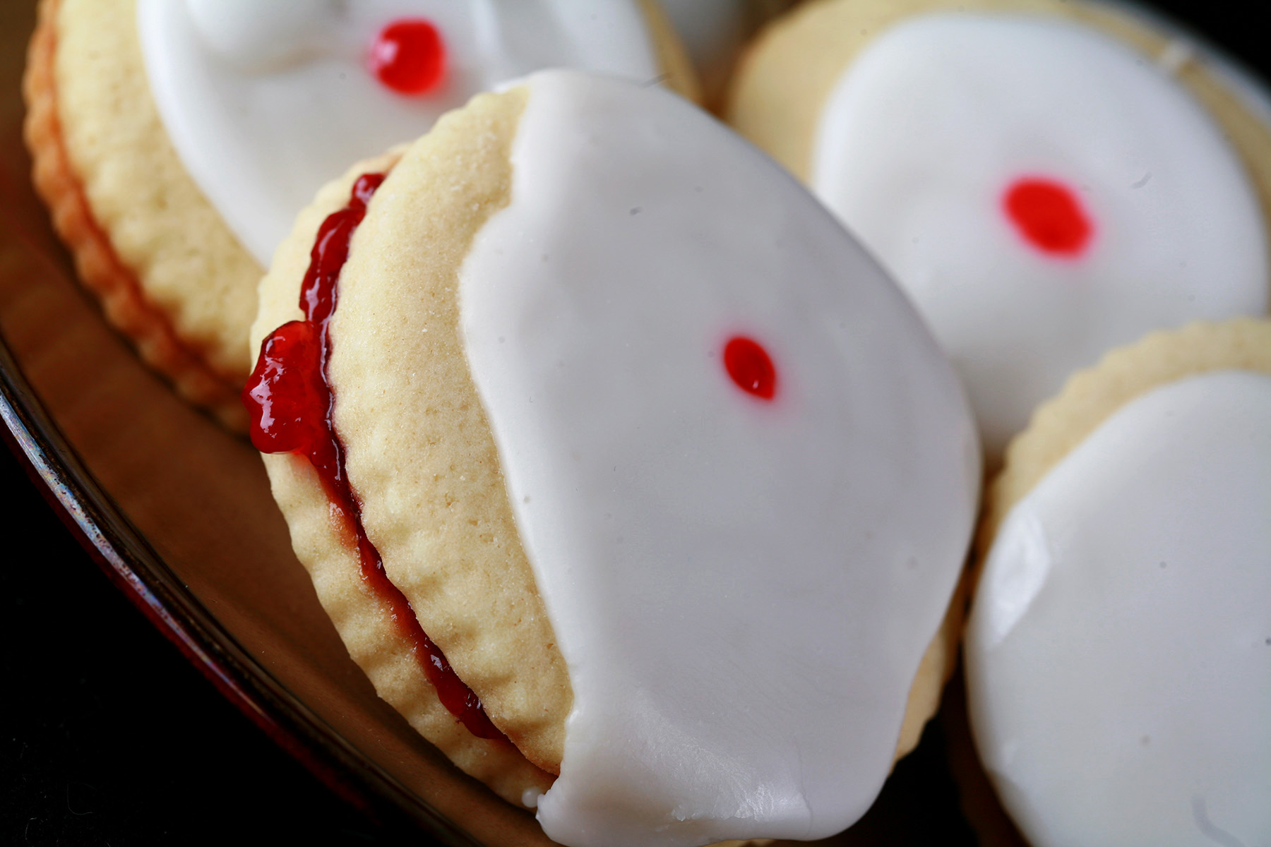 Close up view of a plate of Gluten-Free Empire Biscuits - sandwich cookies filled with raspberry jam, frosted with a white glaze, and finished off with a dot of red gel in the center.