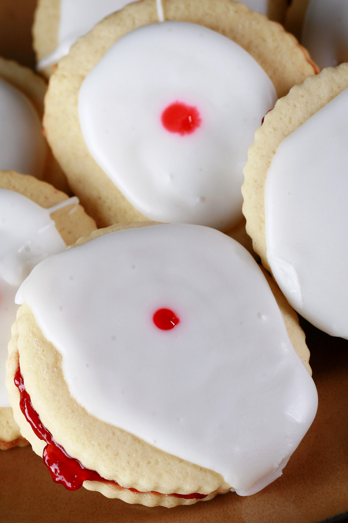 Close up view of a plate of Gluten-Free Empire Biscuits - sandwich cookies filled with raspberry jam, frosted with a white glaze, and finished off with a dot of red gel in the center.