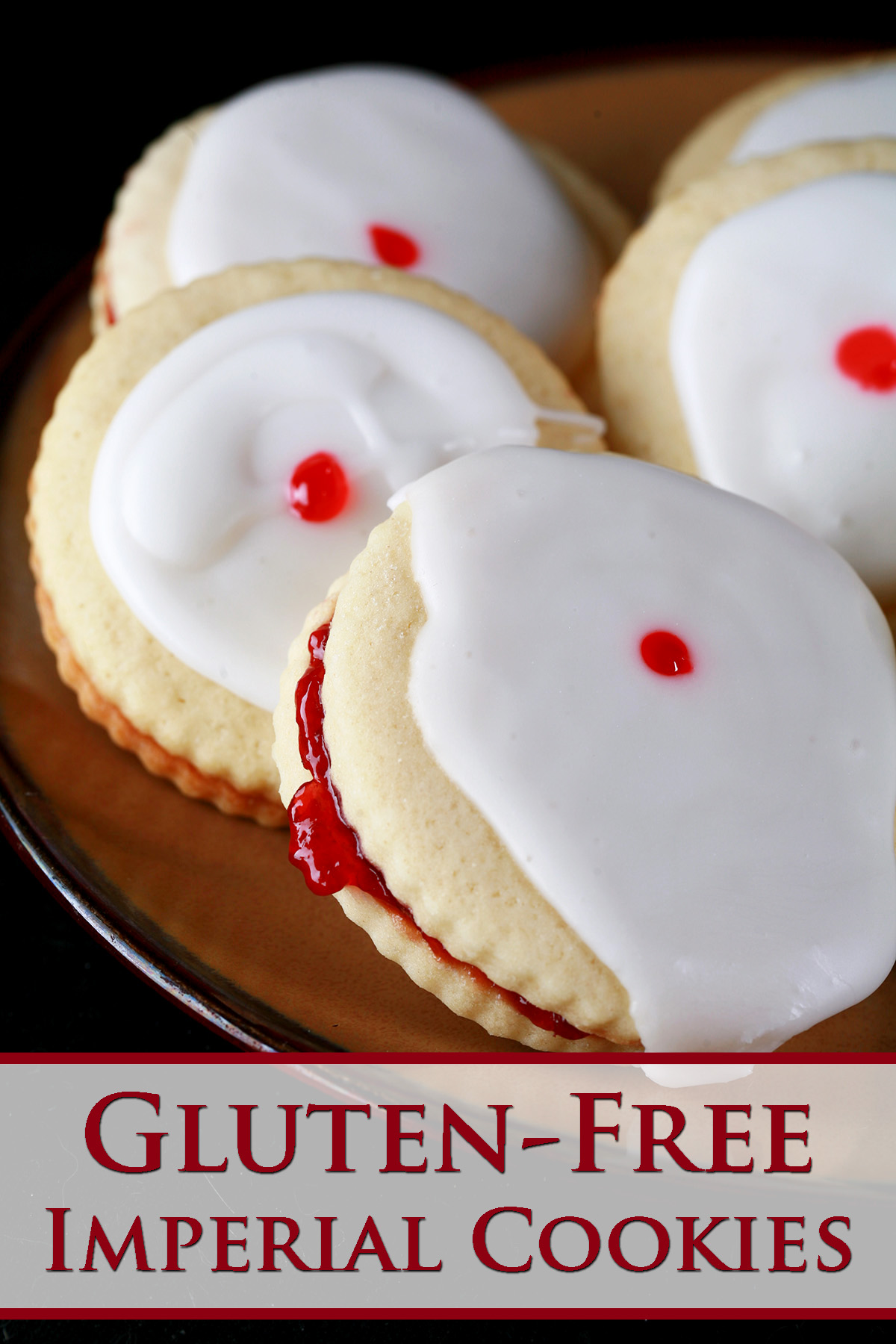 Close up view of a plate of gluten-free imperial cookies: Sandwich cookies filled with raspberry jam, frosted with a white glaze, and finished off with a dot of red gel in the center.