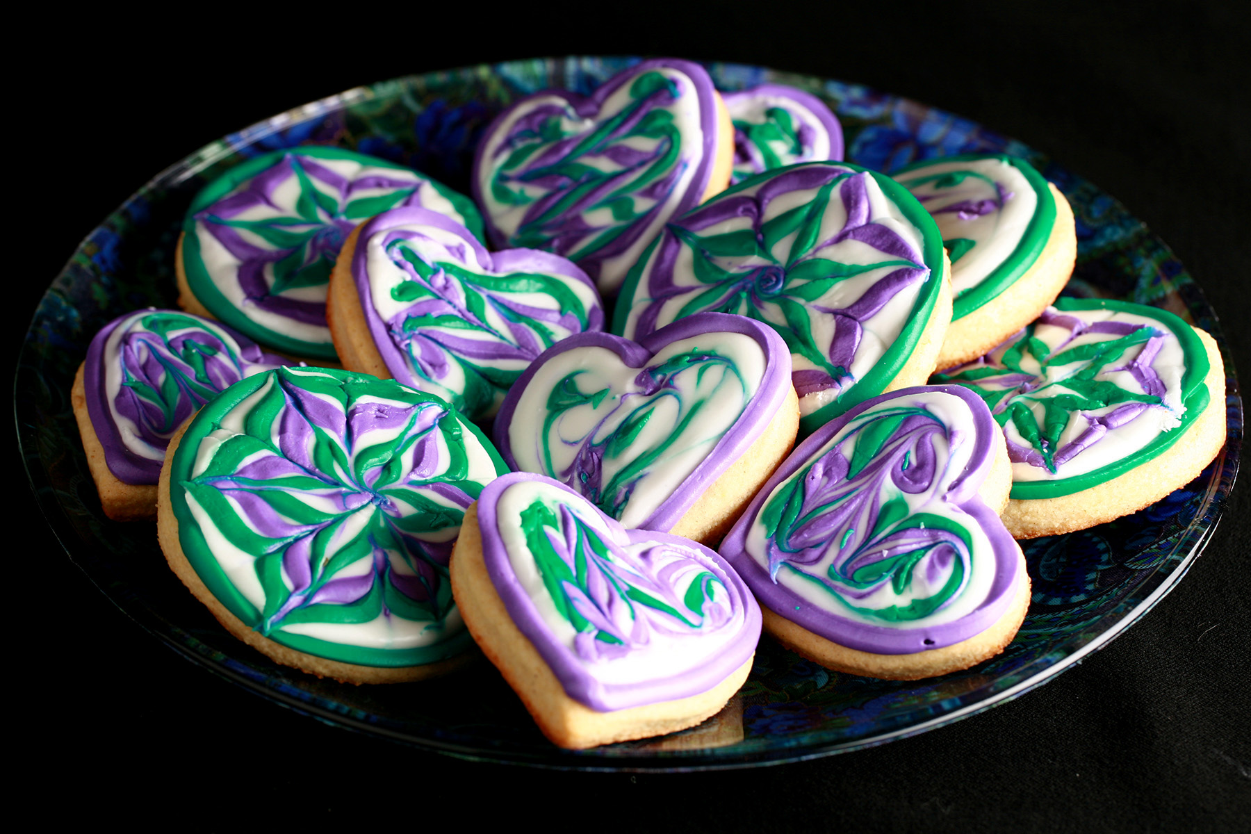 Close up view of a plate of heart shaped gluten-free sugar cookies. They are decorated with swirls of teal and lavender over a base of white frosting.