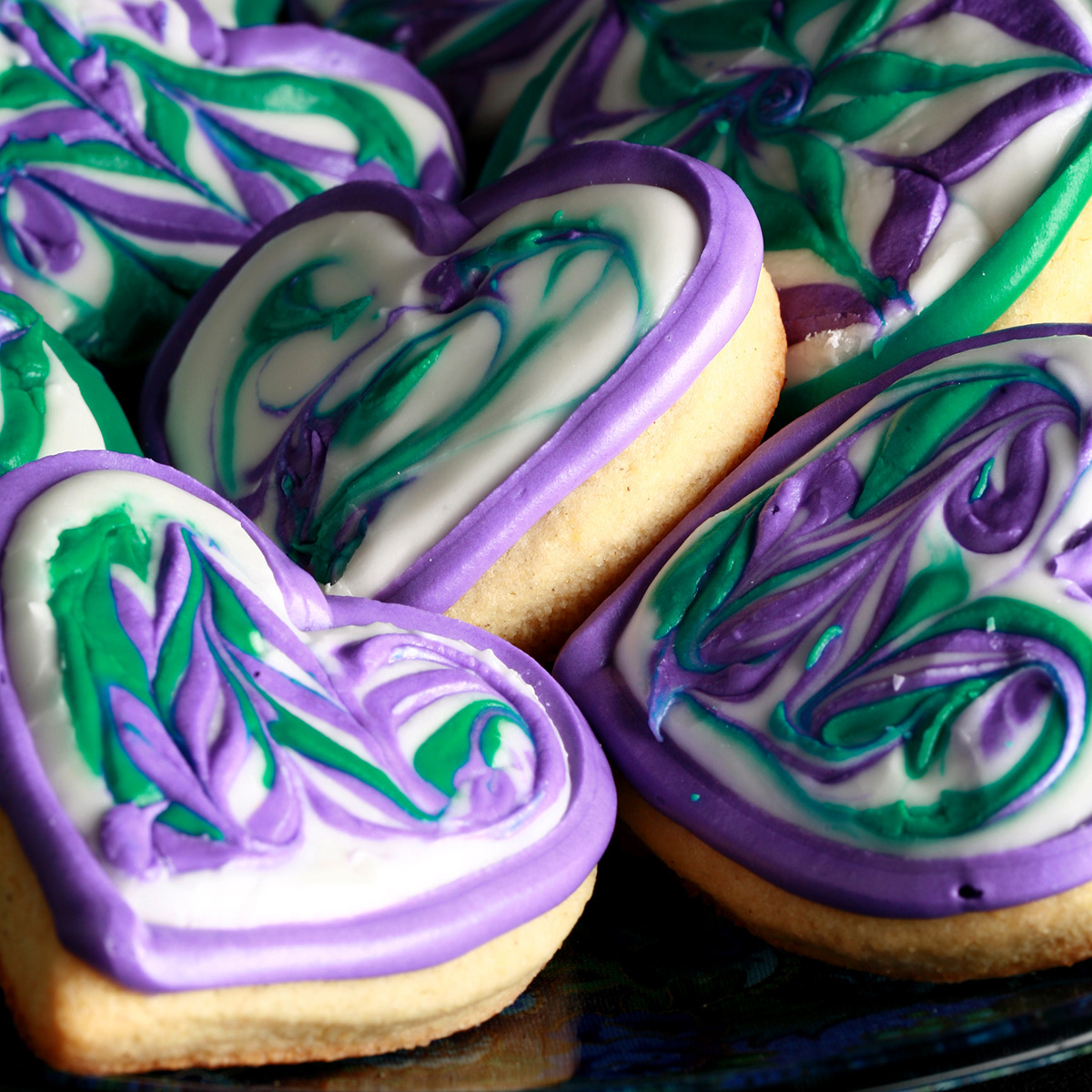 Close up view of a plate of heart shaped gluten-free sugar cookies. They are decorated with swirls of teal and lavender over a base of white frosting