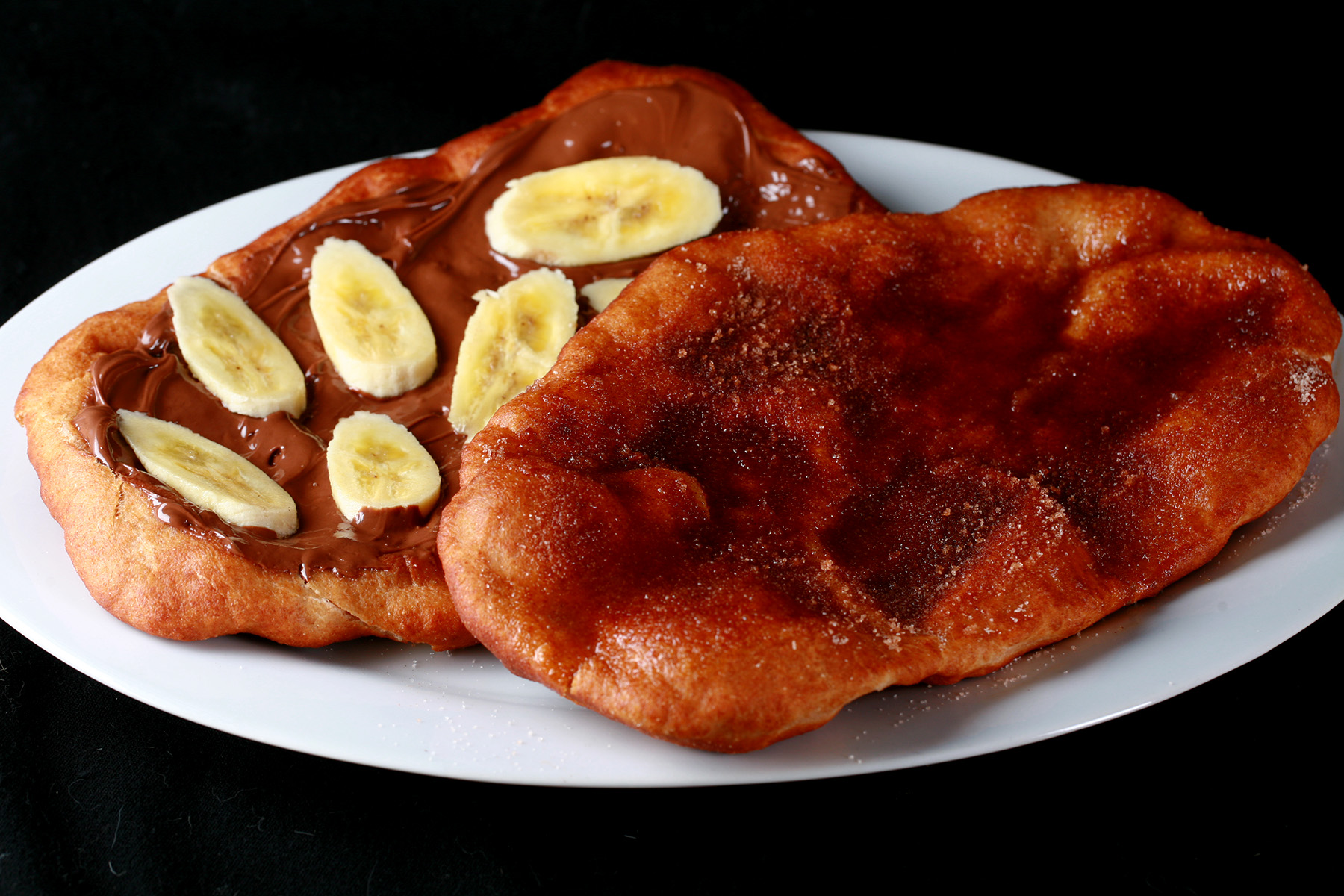 Two gluten free beavertails on a plate. One is cinnamon sugar, the other is nutella and banana.