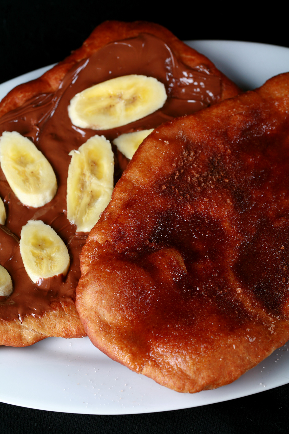 Two gluten-free fried pastries - made from this Gluten-free Beaver Tail recipe - are arranged on a plate. Both are oblong pieces of fried dough. One is topped with cinnamon sugar, the other is spread with Nutella and topped with sliced banana.