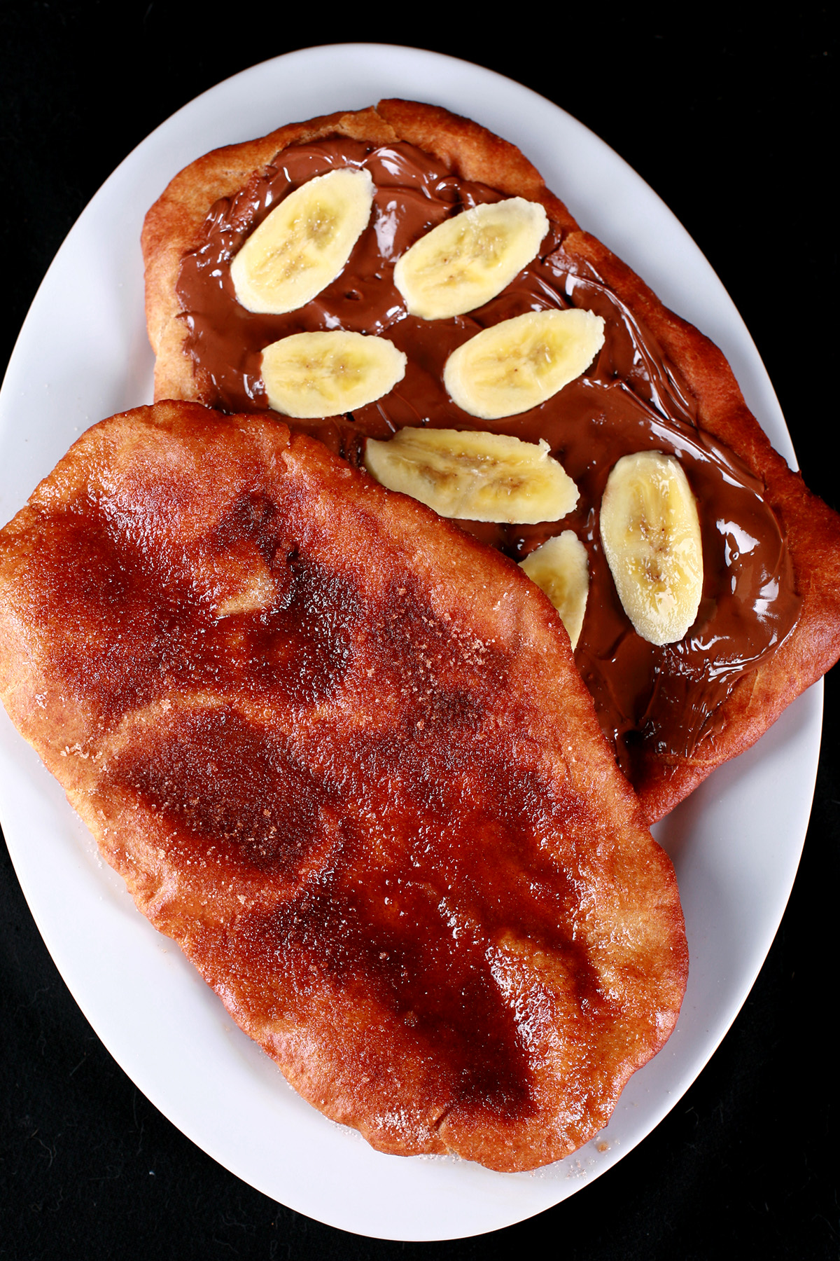 Two gluten-free beaver tail pastries are arranged on a plate. Both are oblong pieces of fried dough. One is topped with cinnamon sugar, the other is spread with Nutella and topped with sliced banana.