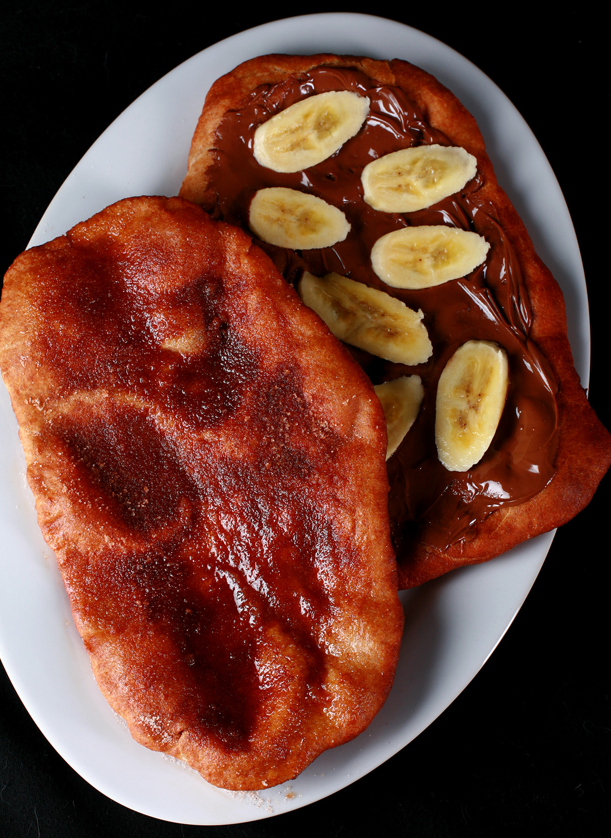 Two gluten-free fried pastries - made from this Gluten-free Beaver Tail recipe - are arranged on a plate. Both are oblong pieces of fried dough. One is topped with cinnamon sugar, the other is spread with Nutella and topped with sliced banana.