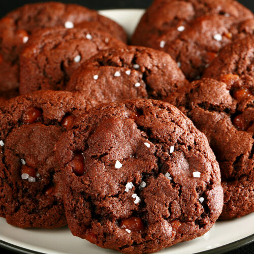 A small white plate, piled with Chewy, Gluten-Free Chocolate Cookies. Bits of caramel are visible within the cookies, and each is topped with a sprinkle of sea salt.