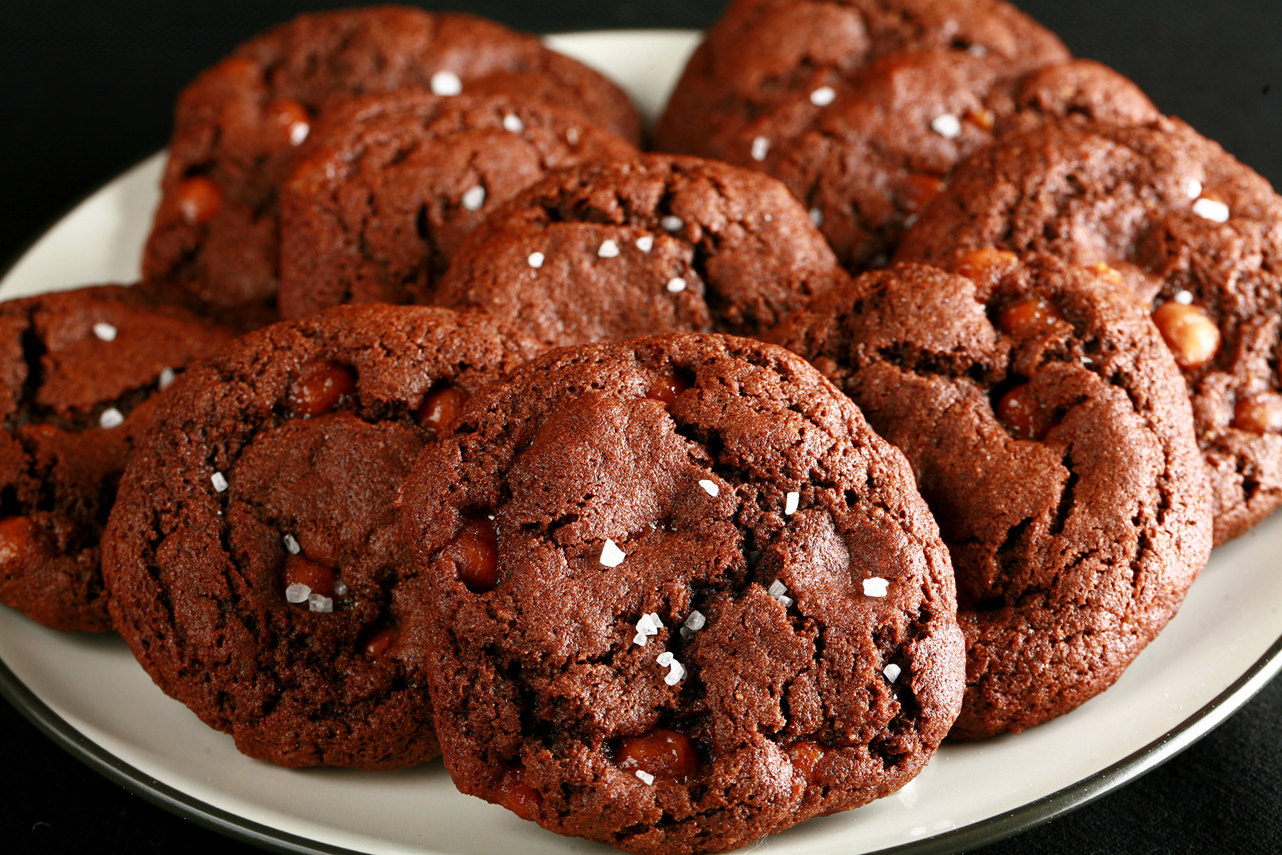 A small white plate, piled with Chewy, Gluten-Free Chocolate Cookies. Bits of caramel are visible within the cookies, and each is topped with a sprinkle of sea salt.