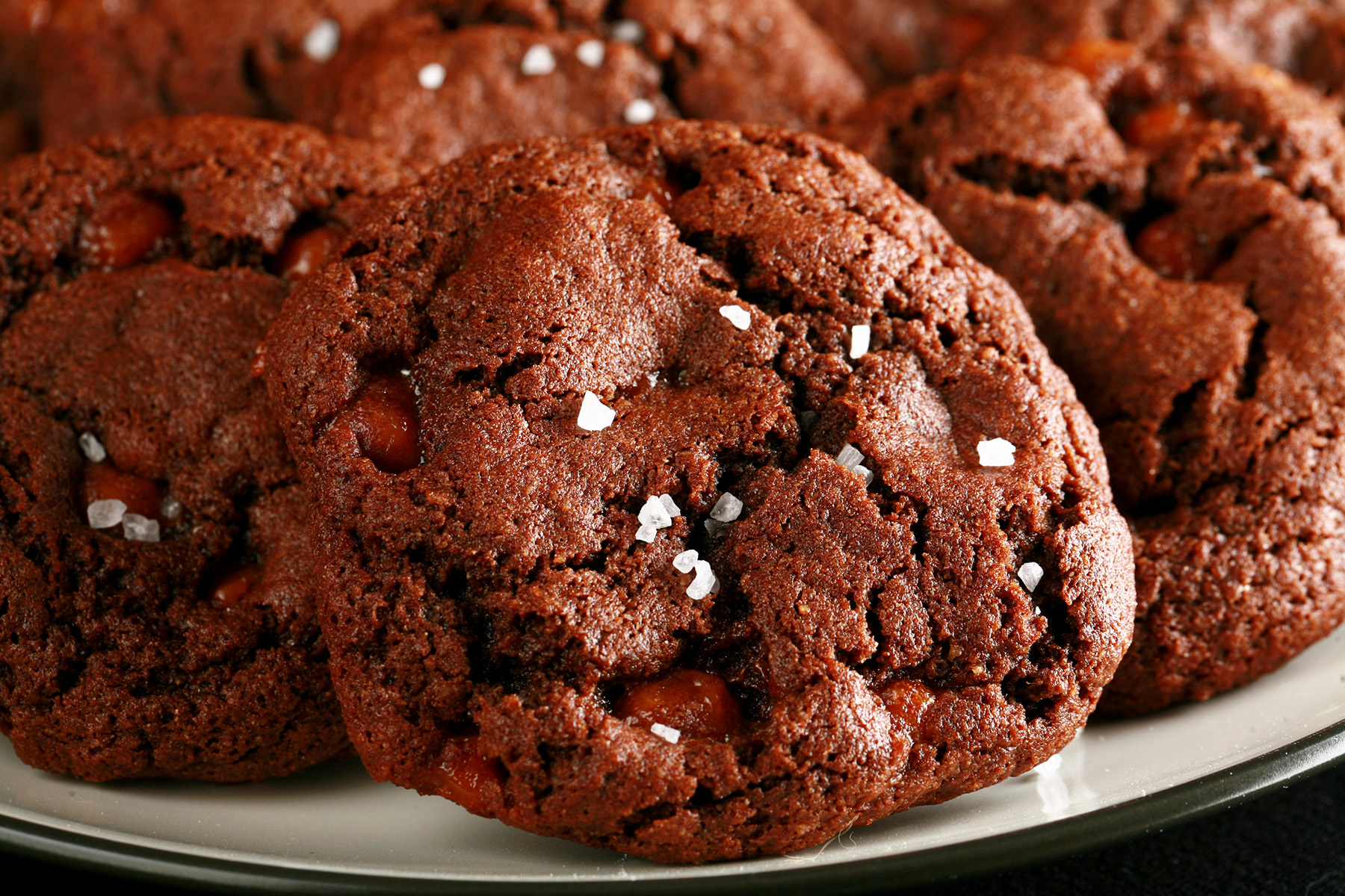 A small white plate, piled with Chewy, Gluten-Free Chocolate Cookies. Bits of caramel are visible within the cookies, and each is topped with a sprinkle of sea salt.