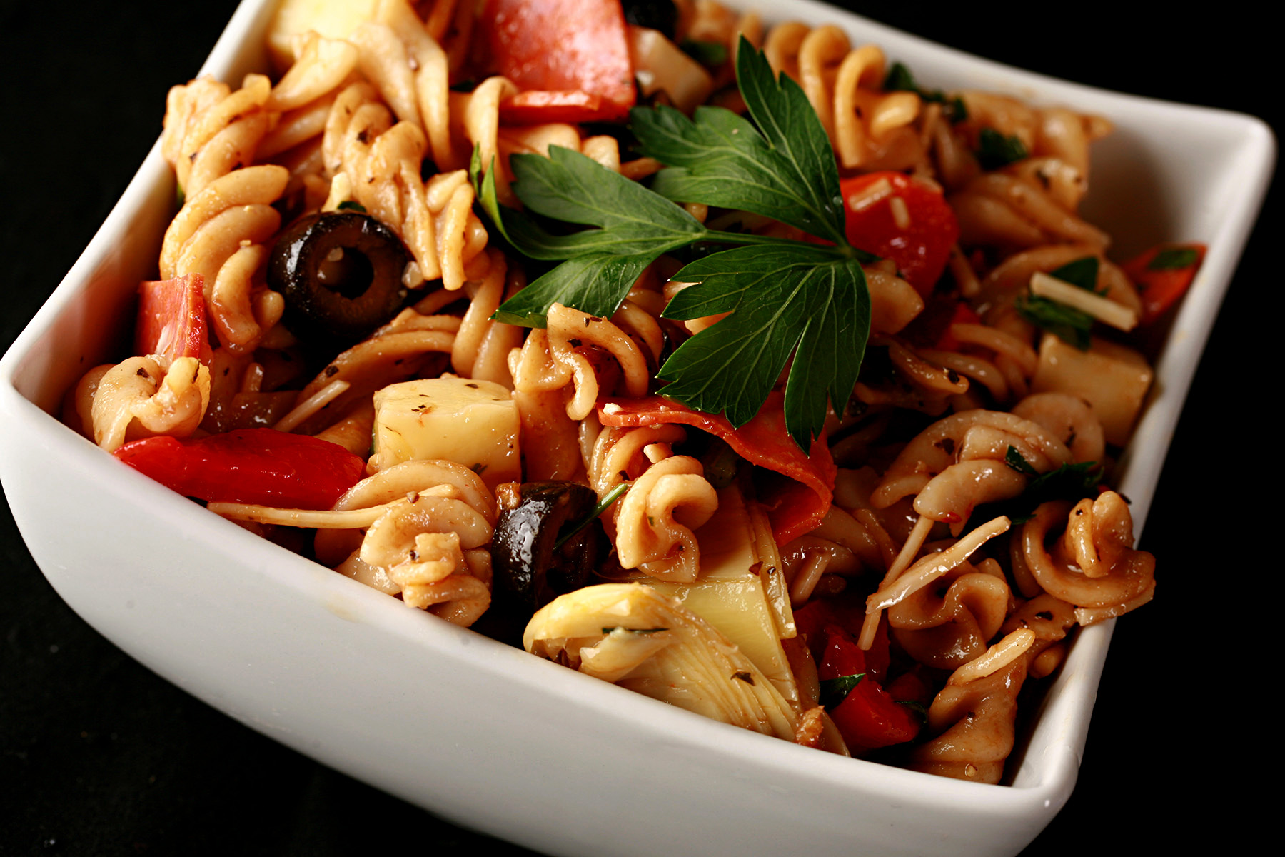 A rich looking pasta salad in a white bowl. Spiral noodles are shown lgthly coated in a thin red sauce, with pepperoni, artichoked, black olives, cheese,  and parsley visible.