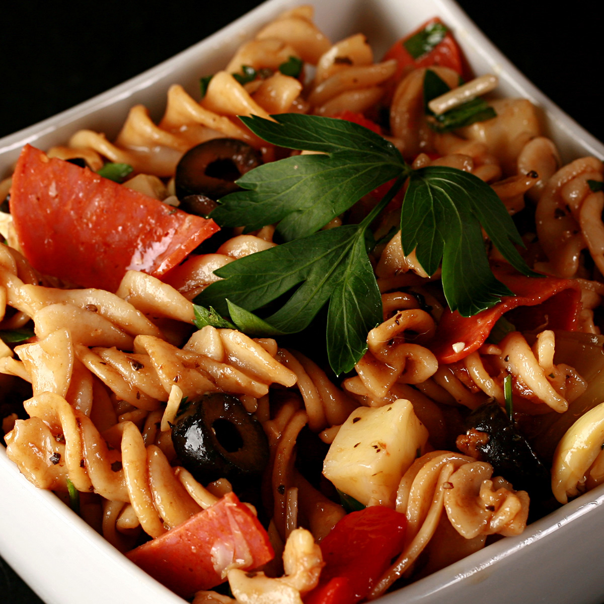 A rich looking pasta salad in a white bowl. Spiral noodles are shown lgthly coated in a thin red sauce, with pepperoni, artichoked, black olives, cheese, and parsley visible.