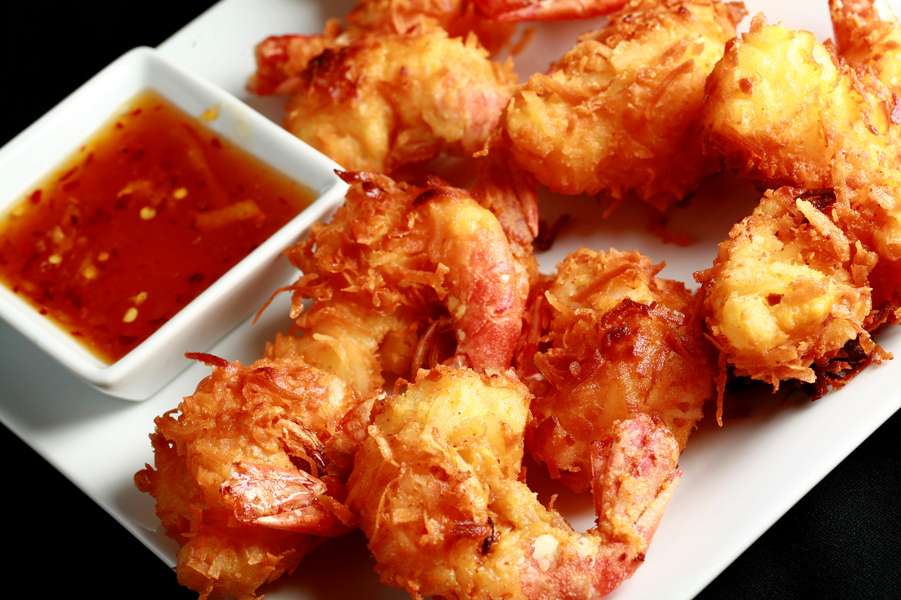 "A white plate is covered with golden brown gluten-free coconut shrimp.  There is a small bowl of spicy ginger-orange sauce on the same plate, as a dip.