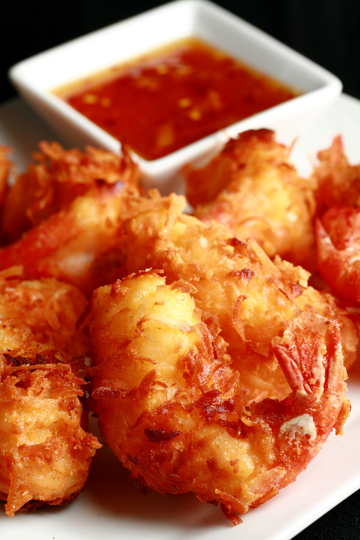 A white plate is covered with golden brown gluten-free coconut shrimp.  There is a small bowl of spicy ginger-orange sauce on the same plate, as a dip.