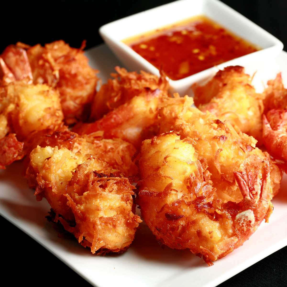 A white plate is covered with golden brown gluten-free coconut shrimp.  There is a small bowl of spicy ginger-orange sauce on the same plate, as a dip.