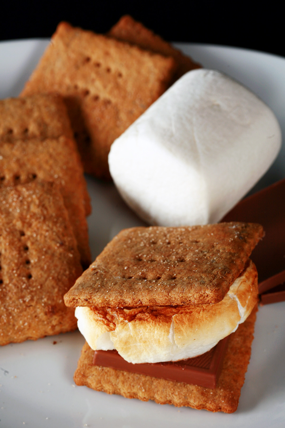 A white plate with rows of square shaped gluten-free graham crackers, a couple large marshmallows, and a piece of chocolate. Two crackers,a piece of chocolate, and a toasted marshmallow have been assembled into a S'more also on the place.