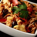 A bowl of Gluten-free pasta salad in a white bowl. Spiral noodles are shown ligthly coated in a thin red sauce, with pepperoni, artichokes, black olives, cheese, and parsley visible.