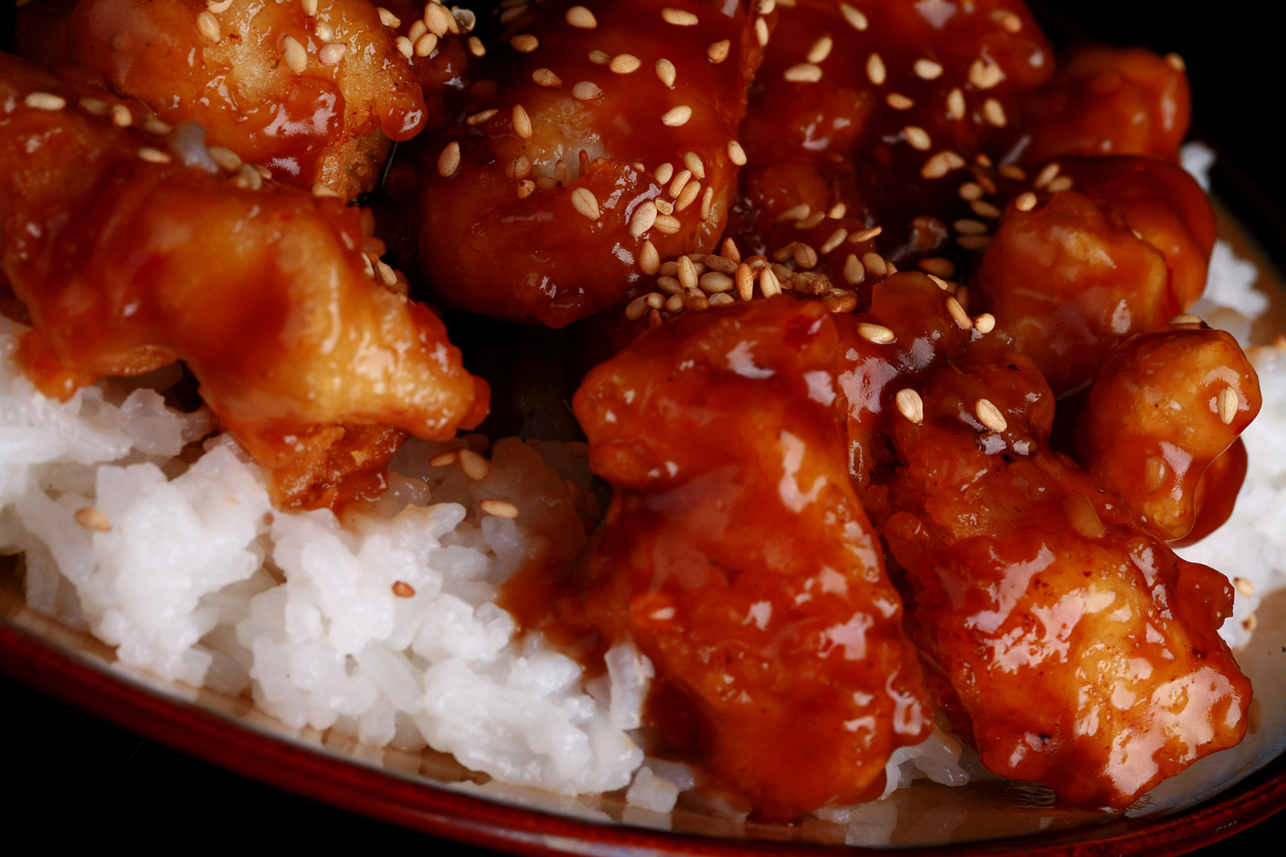 A close up view of a plate of white rice, topped with gluten-free sesame chicken. Chunks of battered and fried chicken in a glossy brown sauce, sprinkled with sesame seeds.