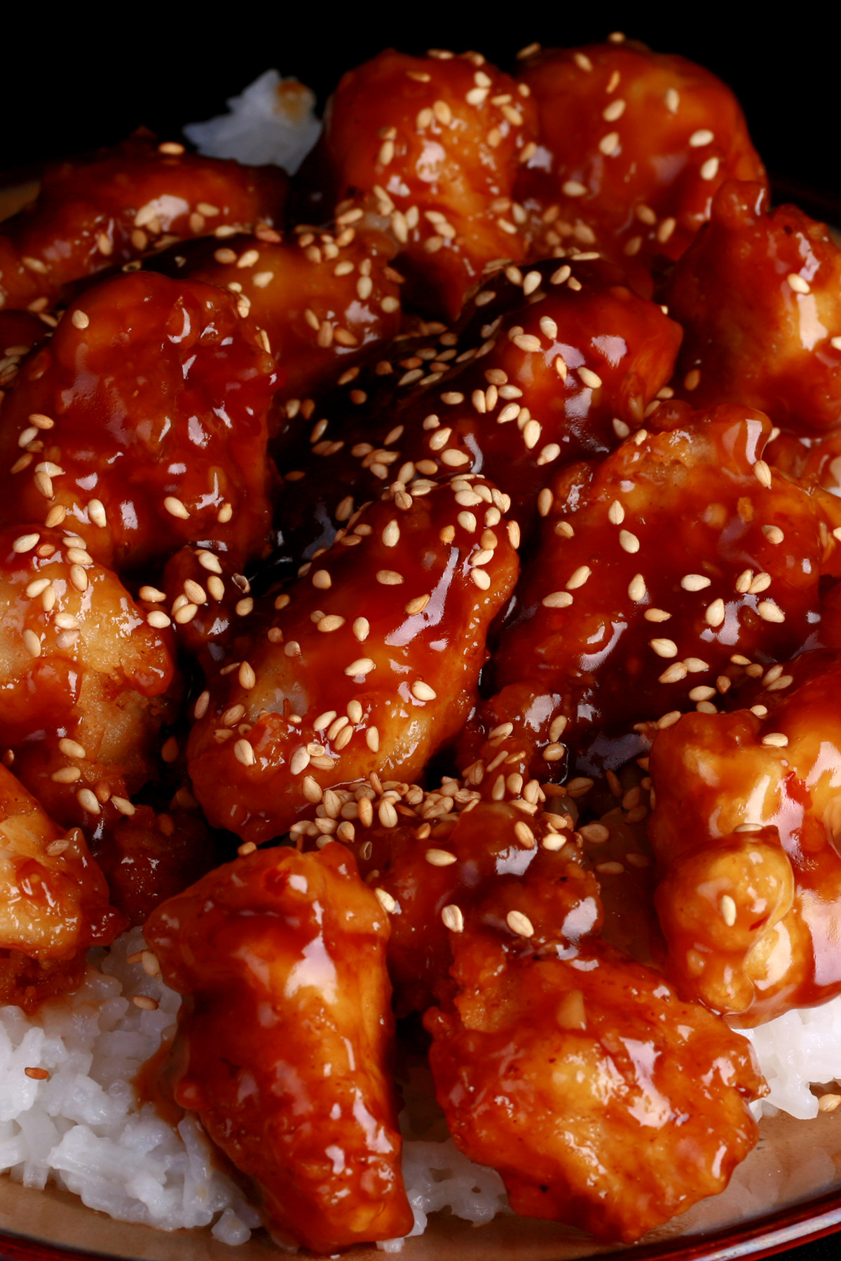 A close up view of a plate of white rice, topped with gluten-free sesame chicken. Chunks of battered and fried chicken in a glossy brown sauce, sprinkled with sesame seeds.