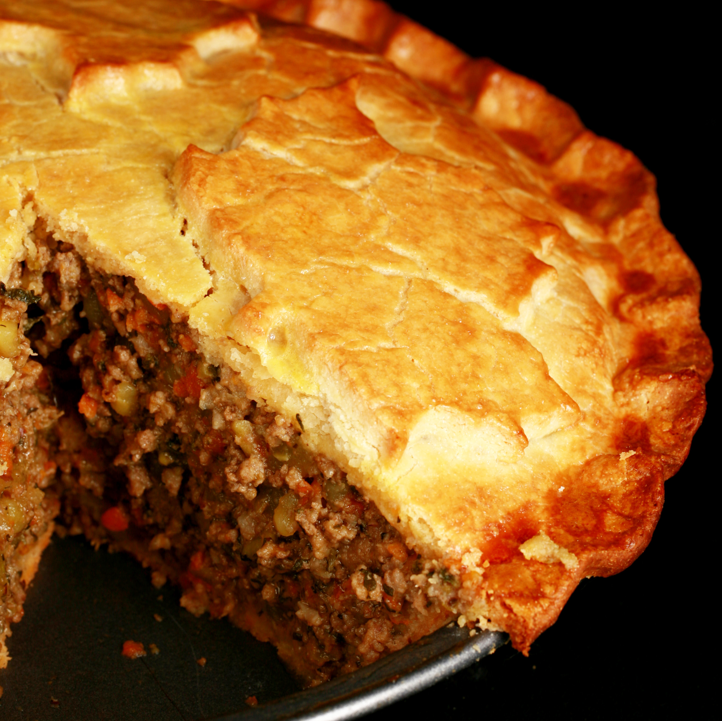 A golden brown gluten-free tourtiere with a maple leaf design on top. There is a section cut out of it, revealing the meat and vegetable filling.