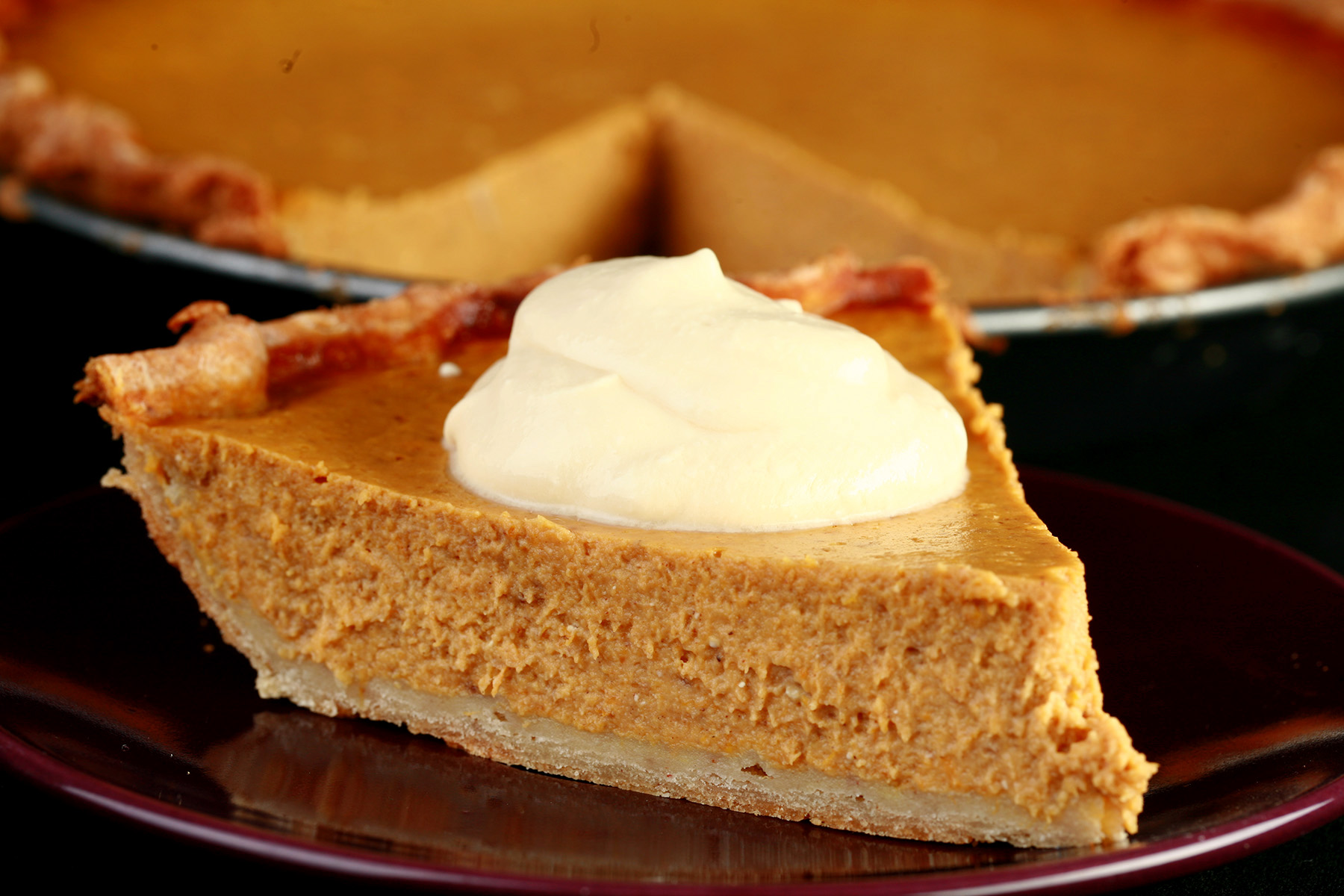 A slice of gluten-free maple pumpkin pie with maple cream, in front of a whole pumpkin pie.
