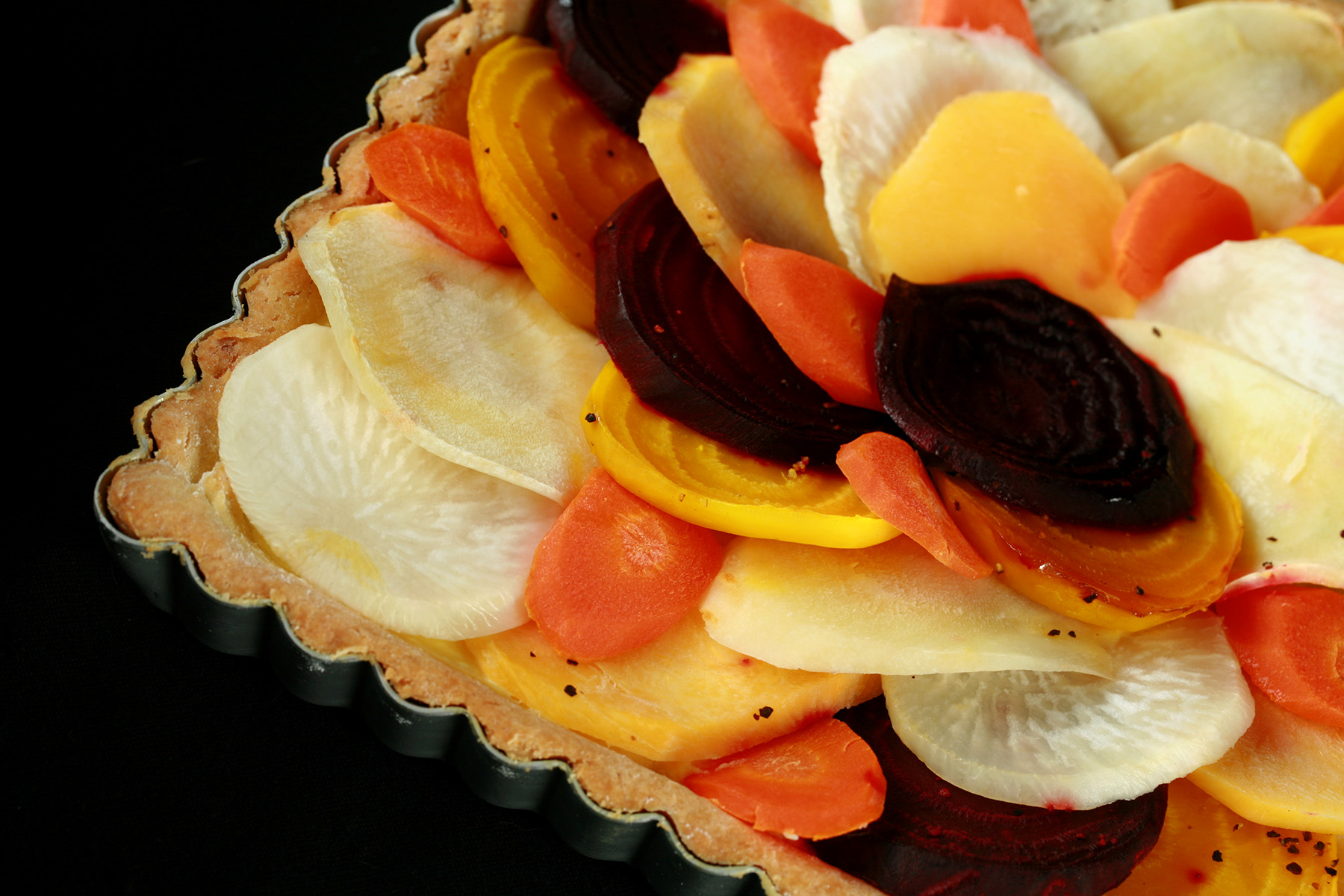 A close up view of a square root tart - a tart cooked in a square pan, with slices of a variety of root veggies arranged on top.