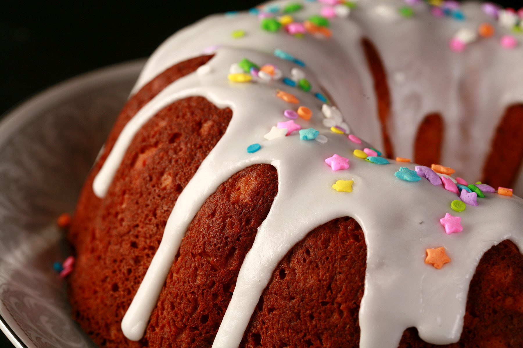 A close up view of a gluten-free paska. It's in the shape of a bundt pan, with dripping white glaze and topped with pastel coloured sprinkles.