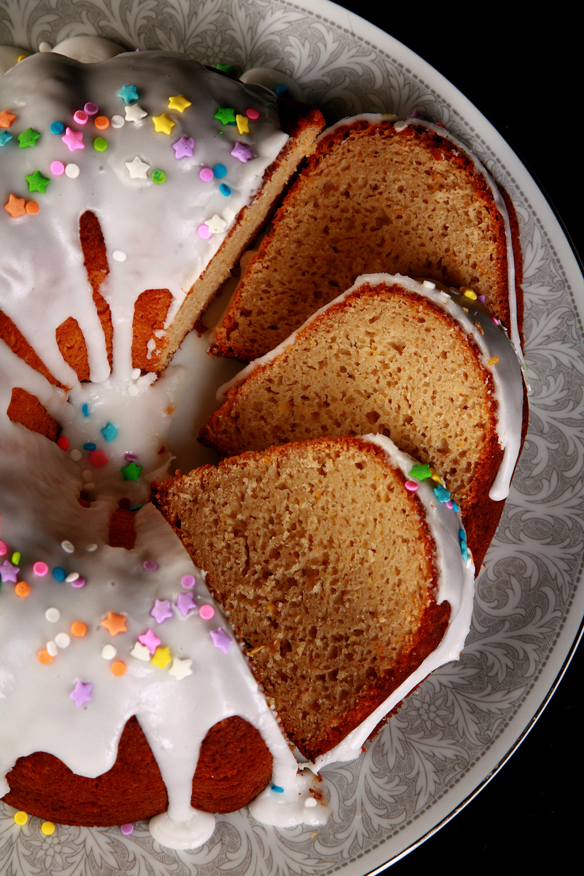 A close up view of a gluten-free paska. It's in the shape of a bundt pan, with dripping white glaze and topped with pastel coloured sprinkles. A section has been sliced, the slices are fanned out on the plate.