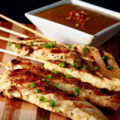 Several skewers of gluten-free chicken satay on a striped cutting board. They are sprinkled with green onions and sitting next to a bowl of peanut sauce.