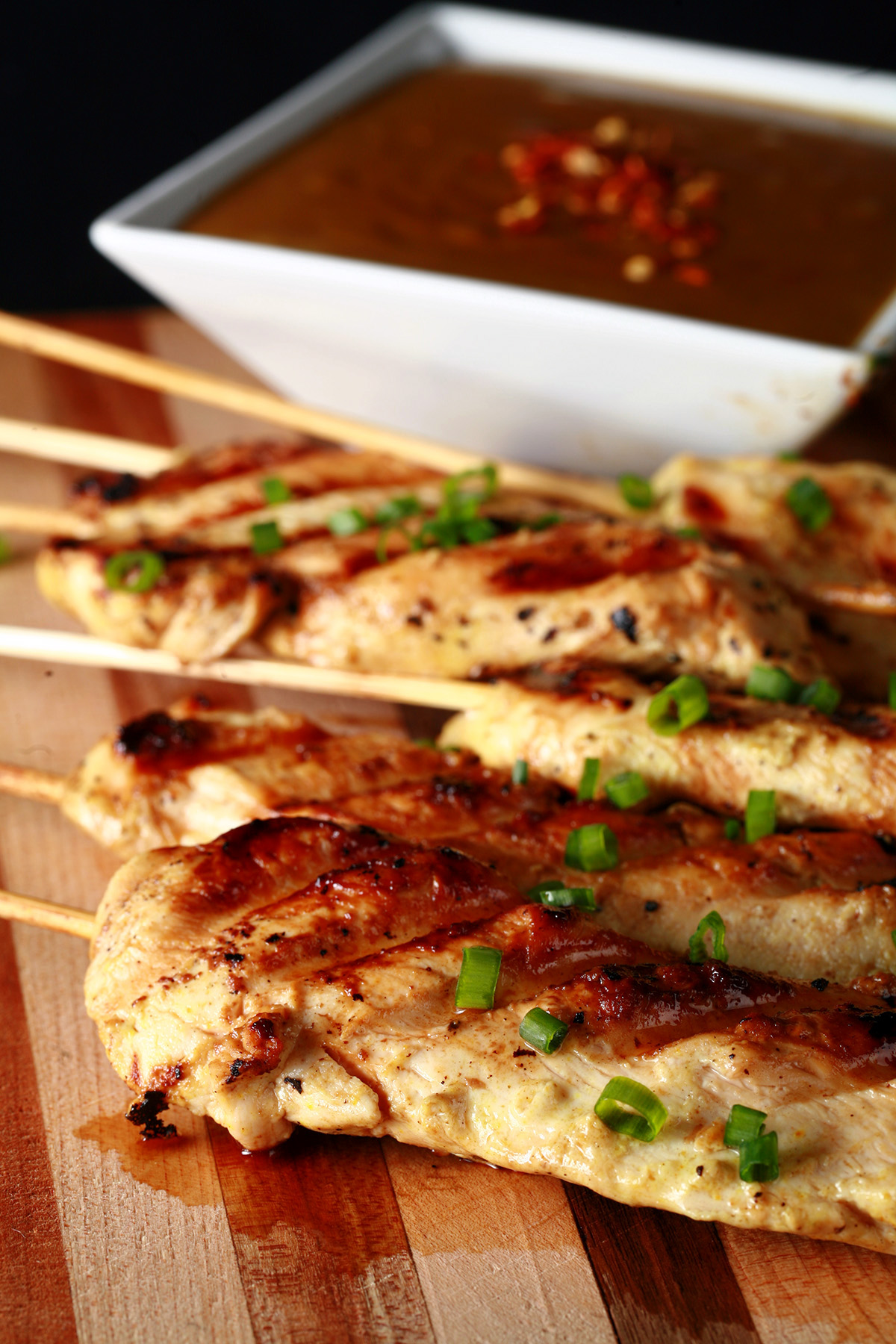 Several skewers of gluten-free chicken satay on a striped cutting board.  They are sprinkled with green onions and sitting next to a bowl of peanut sauce.