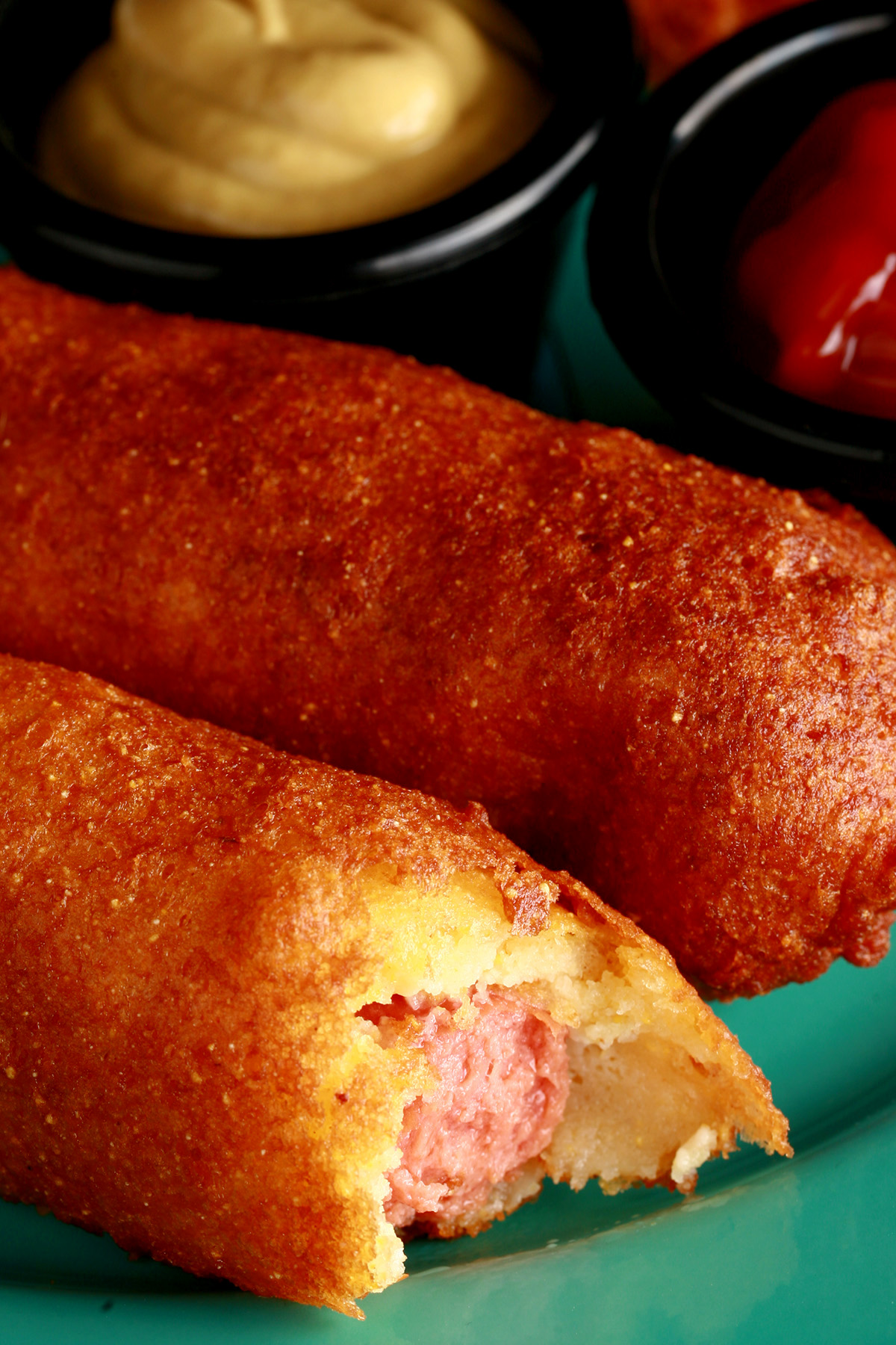 3 gluten-free corndogs on a green plate, with little bowls of ketchup and mustard.
