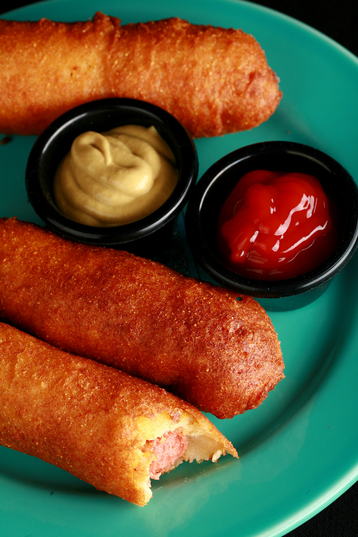 3 gluten-free corn dogs on a green plate, with little bowls of ketchup and mustard.