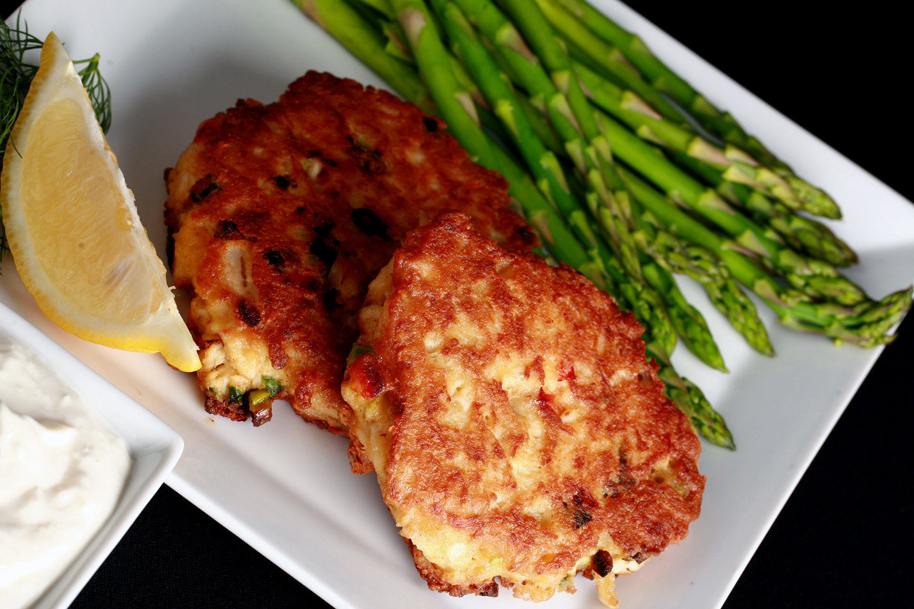 2 Gluten-free Crab Cakes on a plate with asparagus, a lemon slice, and some fresh dill.  There is a small bowl of horseradish sauce next to it.