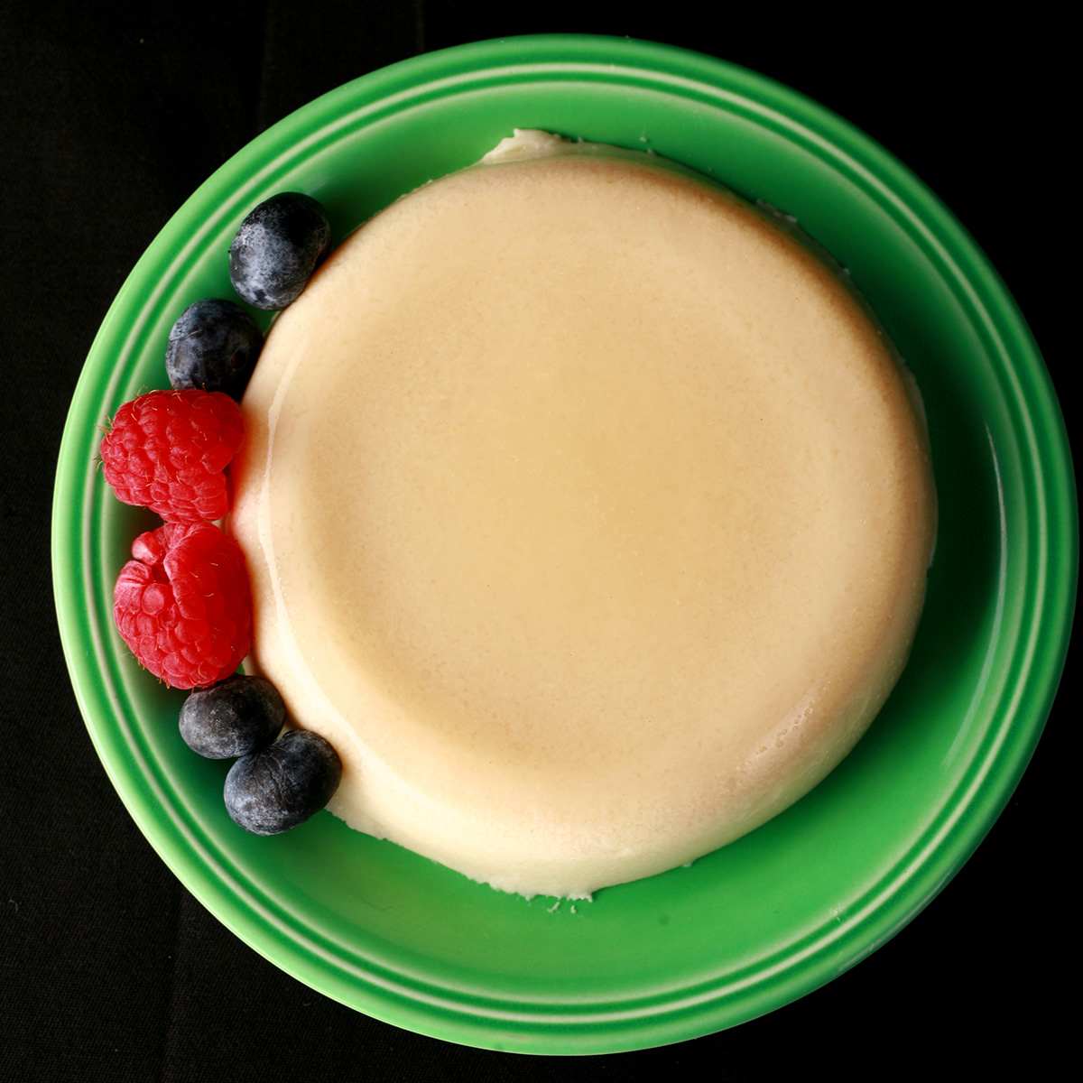 A round serving of calamansi dairy-free panna cotta on a green plate, with raspberries and blueberries next to it.