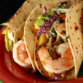A red plate with shrimp tacos. The tacos are made from gluten-free hybrid tortillas, and look like a more yellow version of flour tortillas.