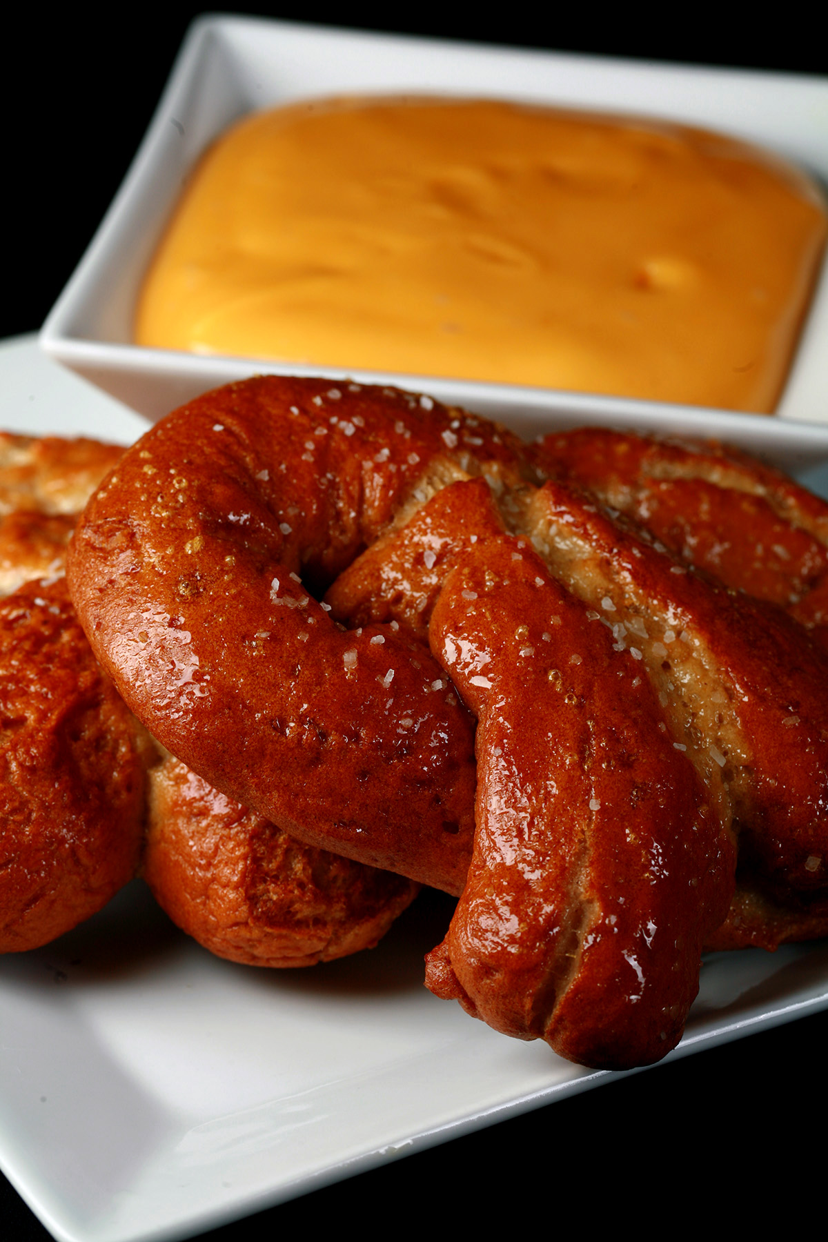 2 gluten-free soft pretzels on a plate, beside a bowl of mustard-cheddar dip.
