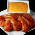 2 gluten-free soft pretzels on a plate, beside a bowl of mustard-cheddar dip.
