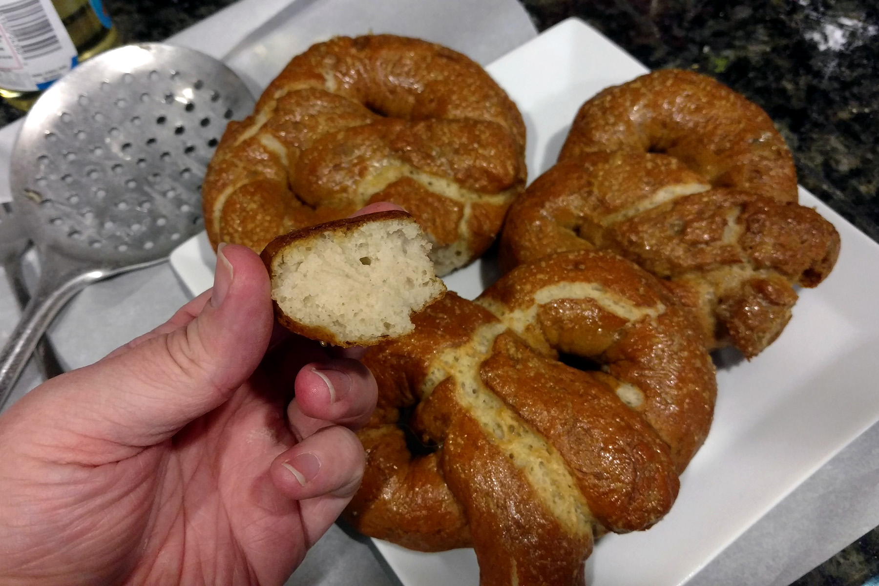 3 gluten free pretzels on a plate, and a hand holding a piece of soft pretzel in the foreground.
