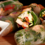 Several mango-shrimp spring rolls on a cutting board, along with a small bowl of peanut sauce.