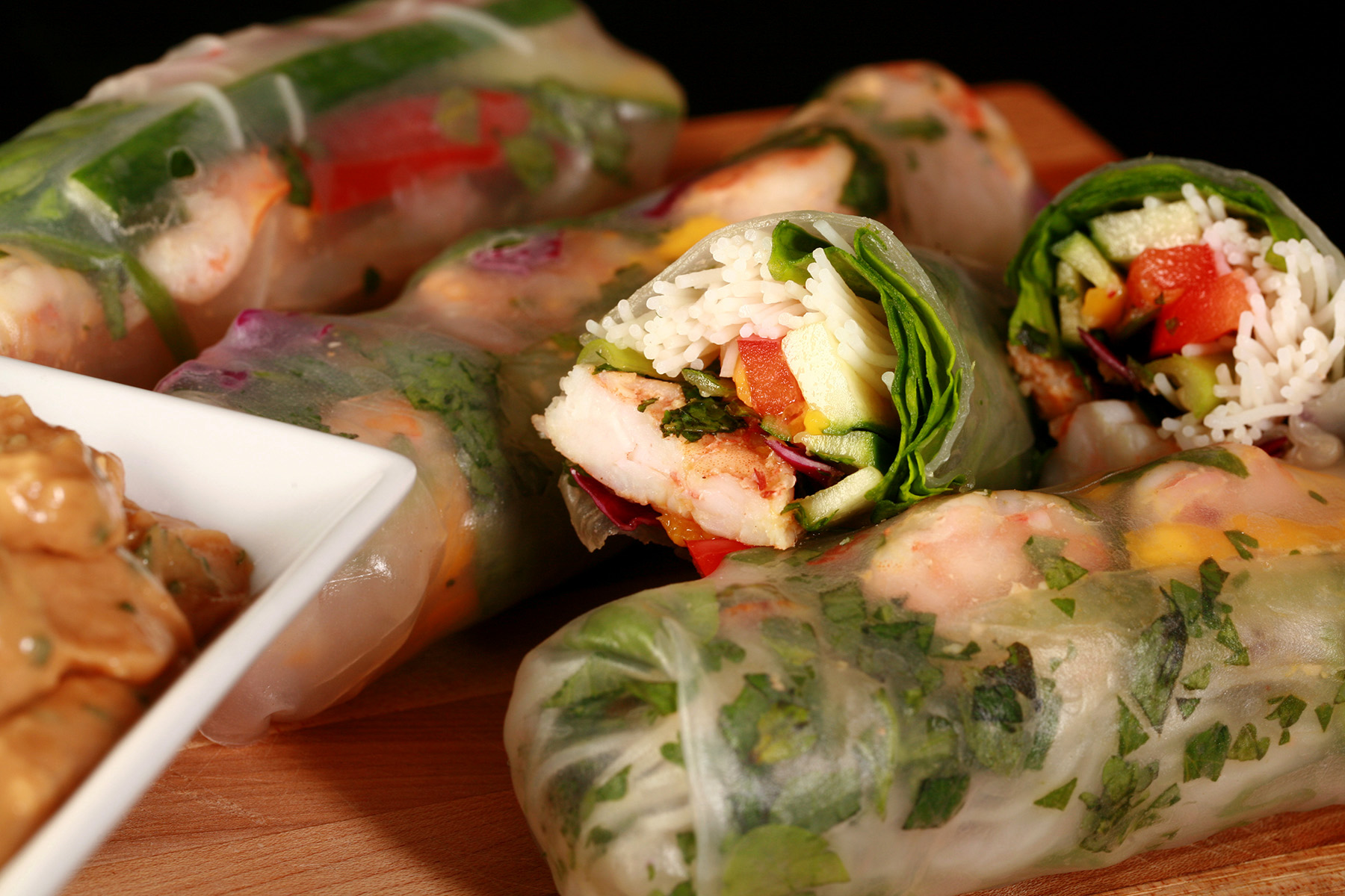 Several mango-shrimp spring rolls on a cutting board, along with a small bowl of peanut sauce.