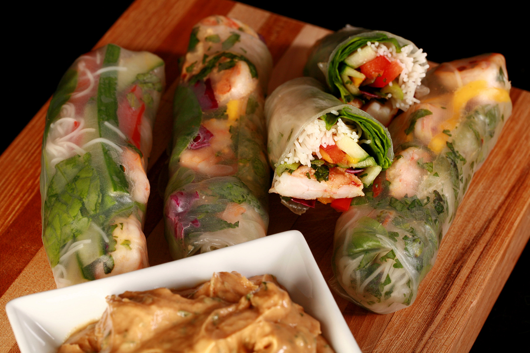 Several mango-shrimp spring rolls on a cutting board, along with a small bowl of peanut sauce.