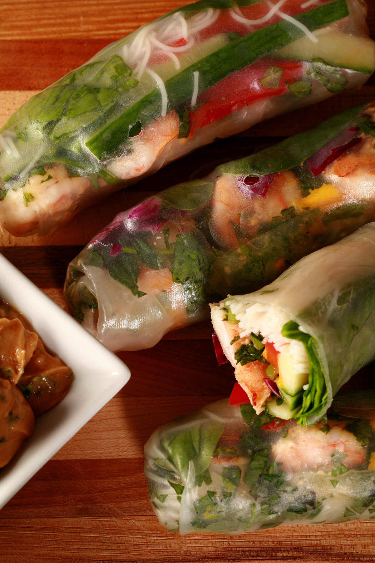 Several mango-shrimp spring rolls on a cutting board, along with a small bowl of peanut sauce.