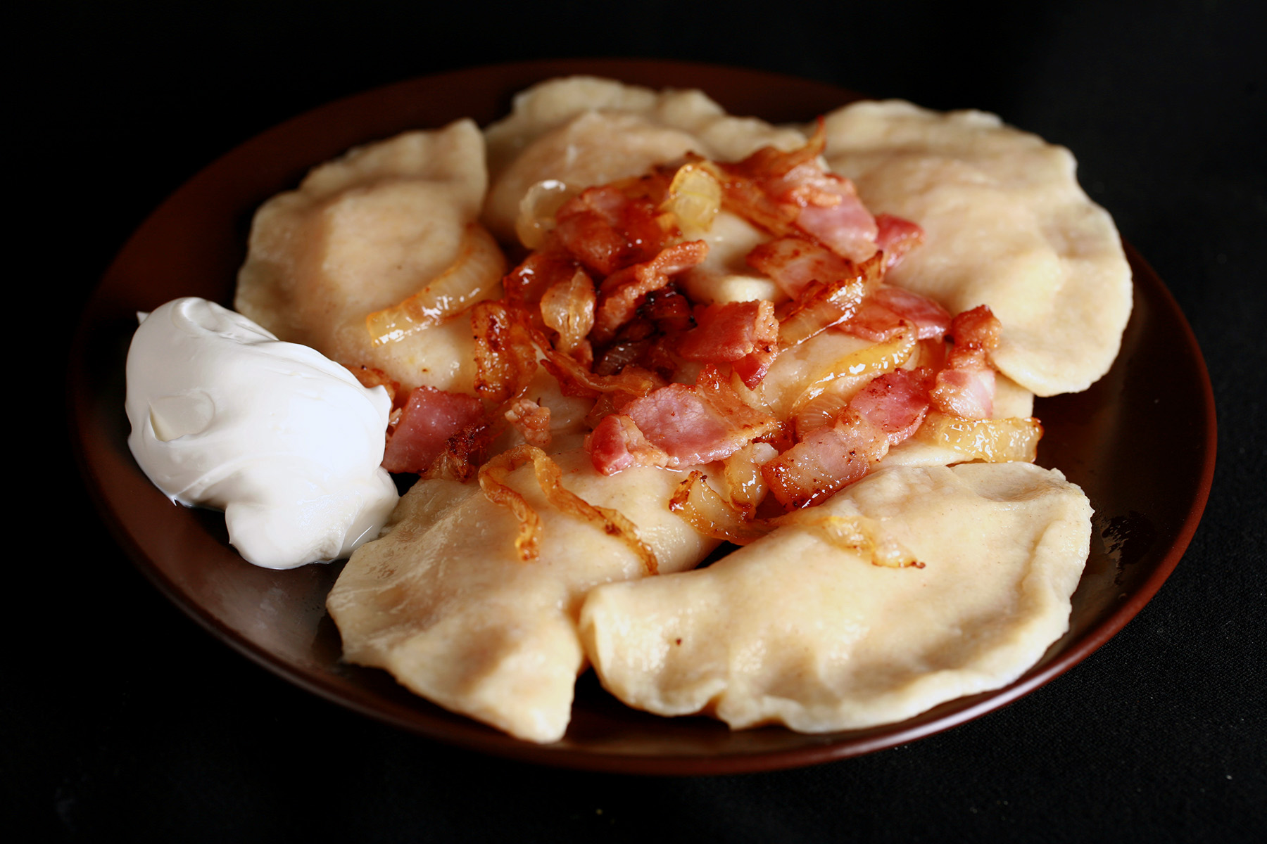 A plate of gluten-free perogies, topped with bacon, fried onions, and sour cream.
