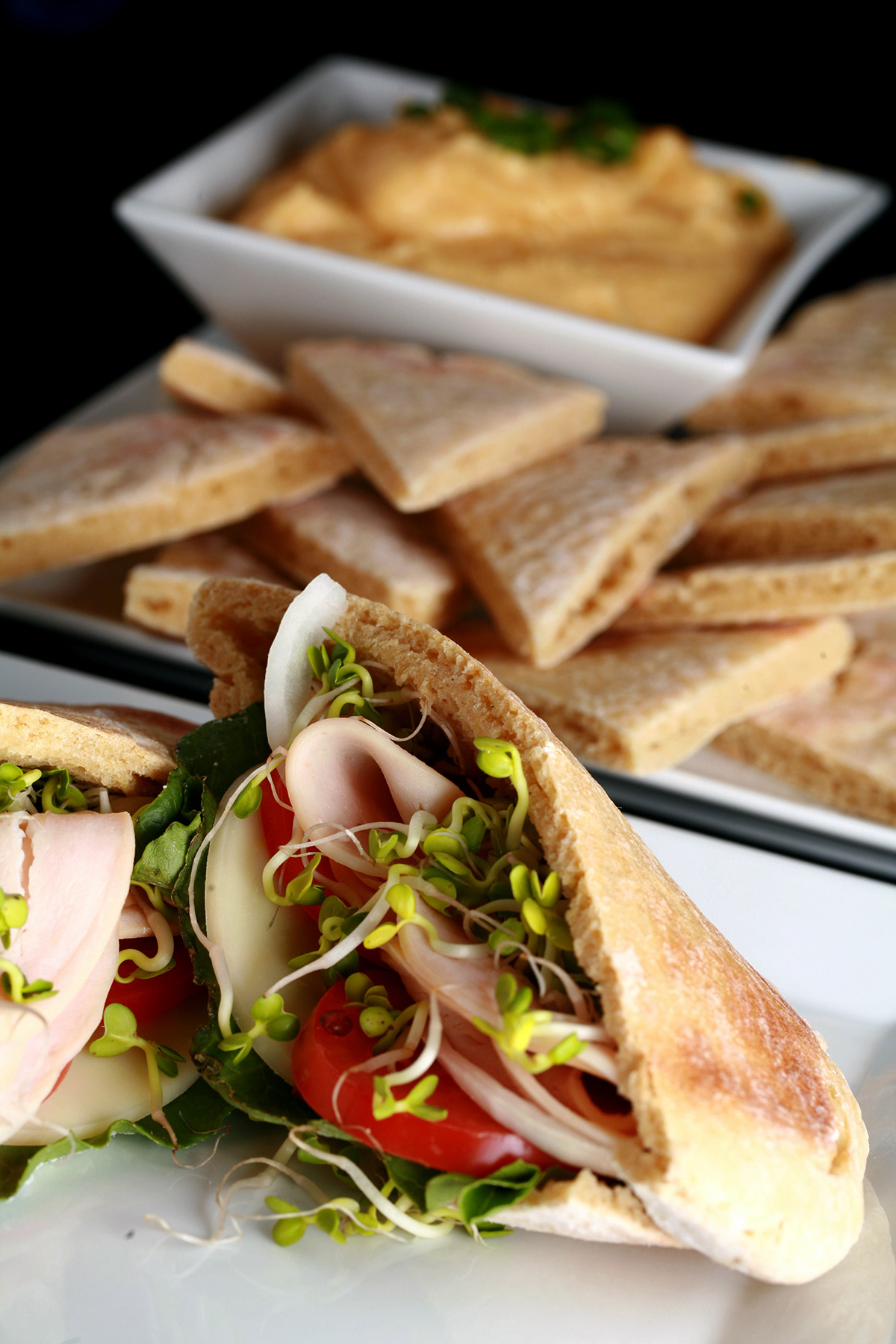 A sandwich made from a gluten free pita pocket, in front of a plate of gluten free pita bread wedges and hummus.