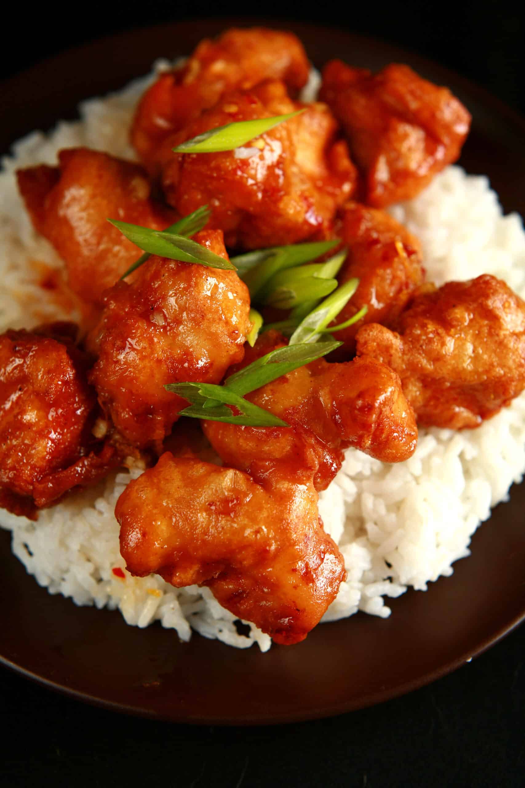 A big helping of gluten free orange chicken: Chunks of battered and deep fried chicken coated in a glossy orange sauce, garnished with sliced green onions, and served over a bed of rice.