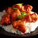 A big helping of gluten-free orange chicken: Chunks of battered and deep fried chicken coated in a glossy orange sauce, garnished with sliced green onions, and served over a bed of rice.