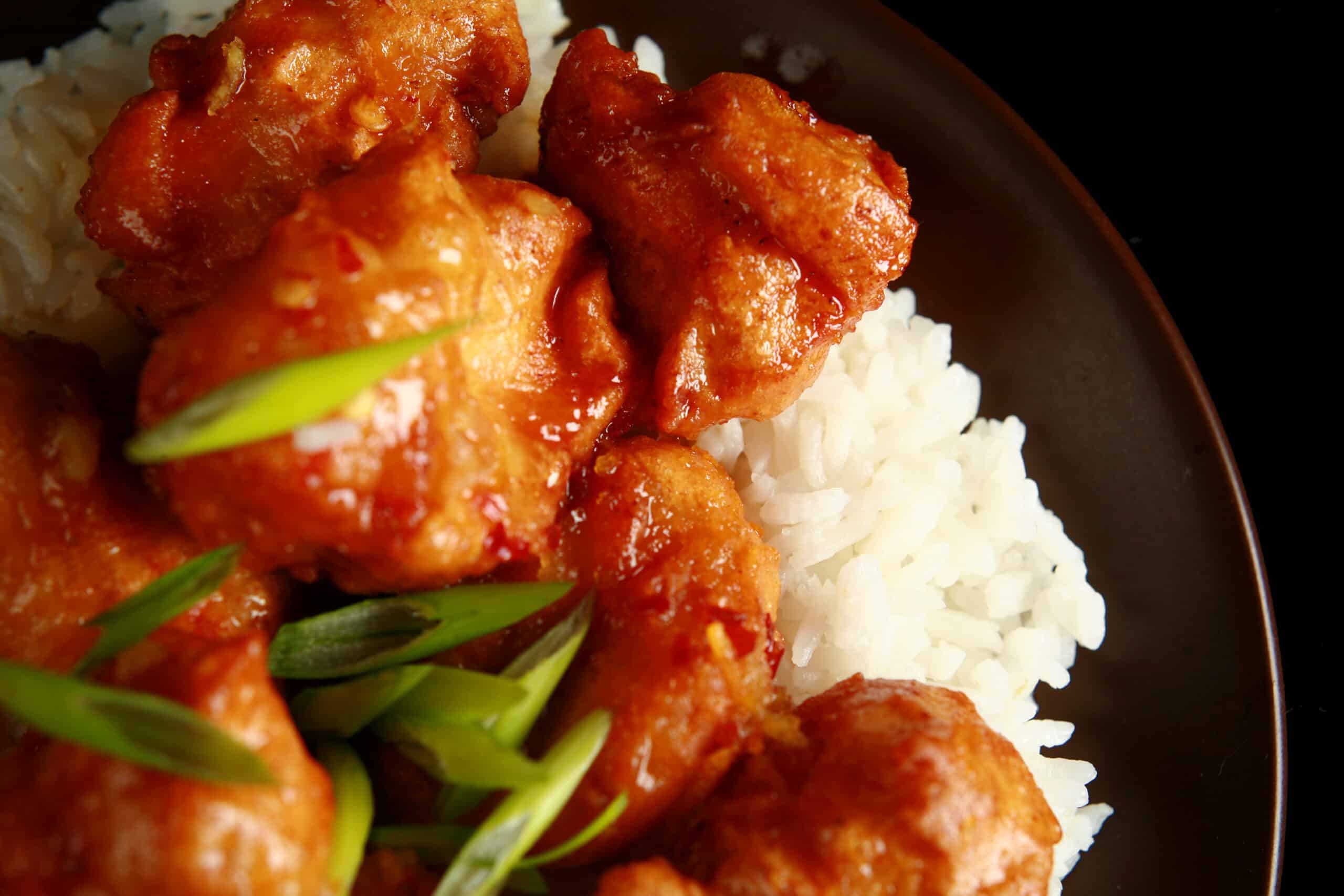 A big helping of gluten free orange chicken: Chunks of battered and deep fried chicken coated in a glossy orange sauce, garnished with sliced green onions, and served over a bed of rice.