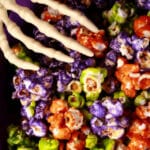 A bowl of Halloween popcorn - orange, purple, and lime green glazed popcorn, mixed together. A pair of skeleton hand serving spoons are visible.