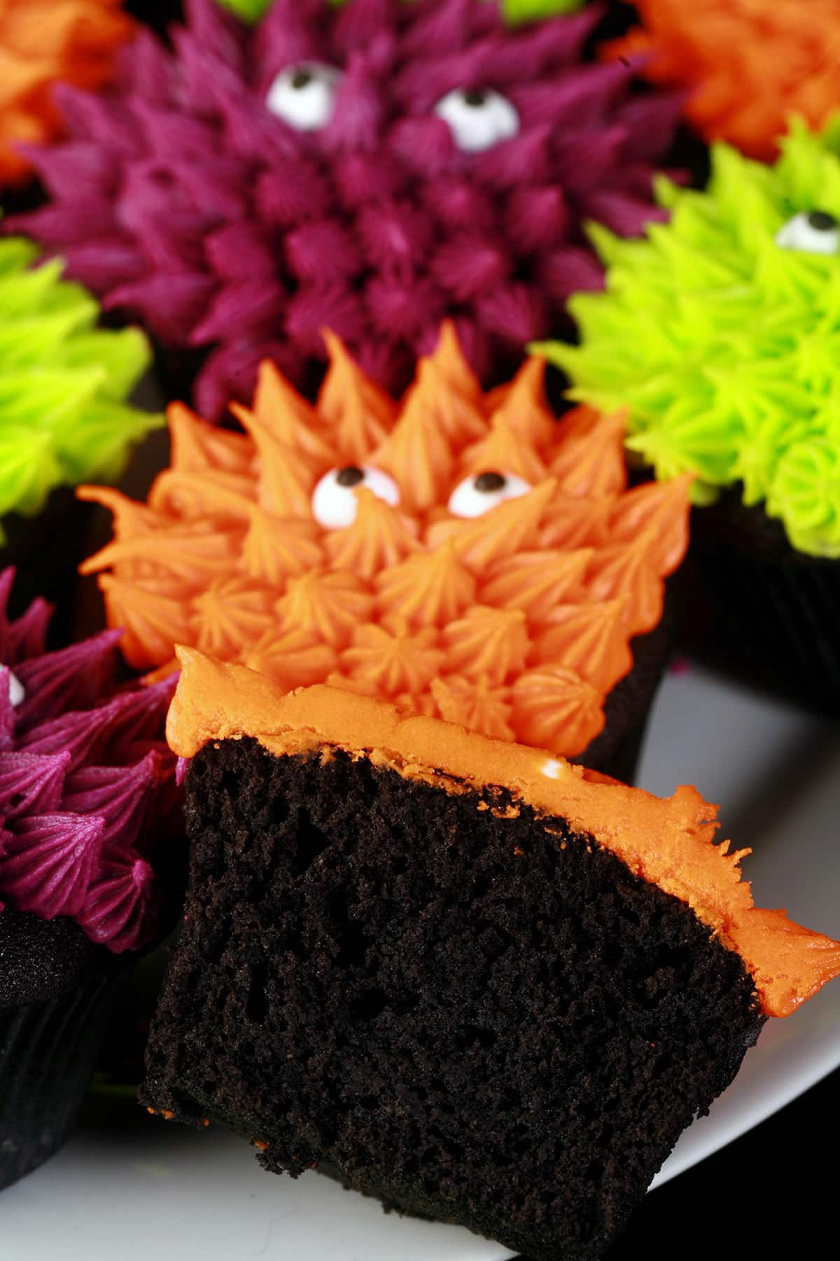 A plate fo gluten free halloween cupcakes, decorated to look like colourful monsters. A split gluten free black velvet cupcake is in the foreground.
