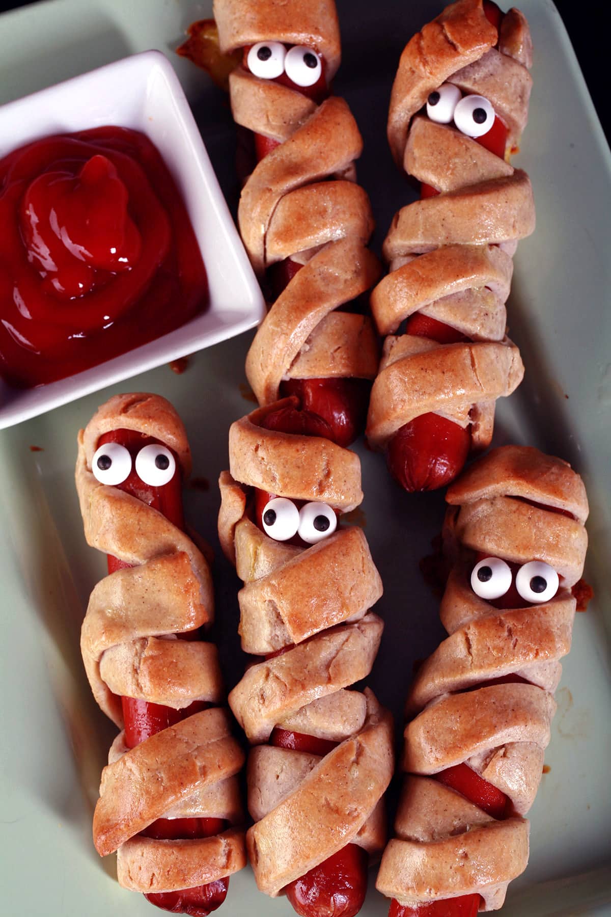 Several gluten-free mummy dogs on a plate. Each have 2 candy eyes.